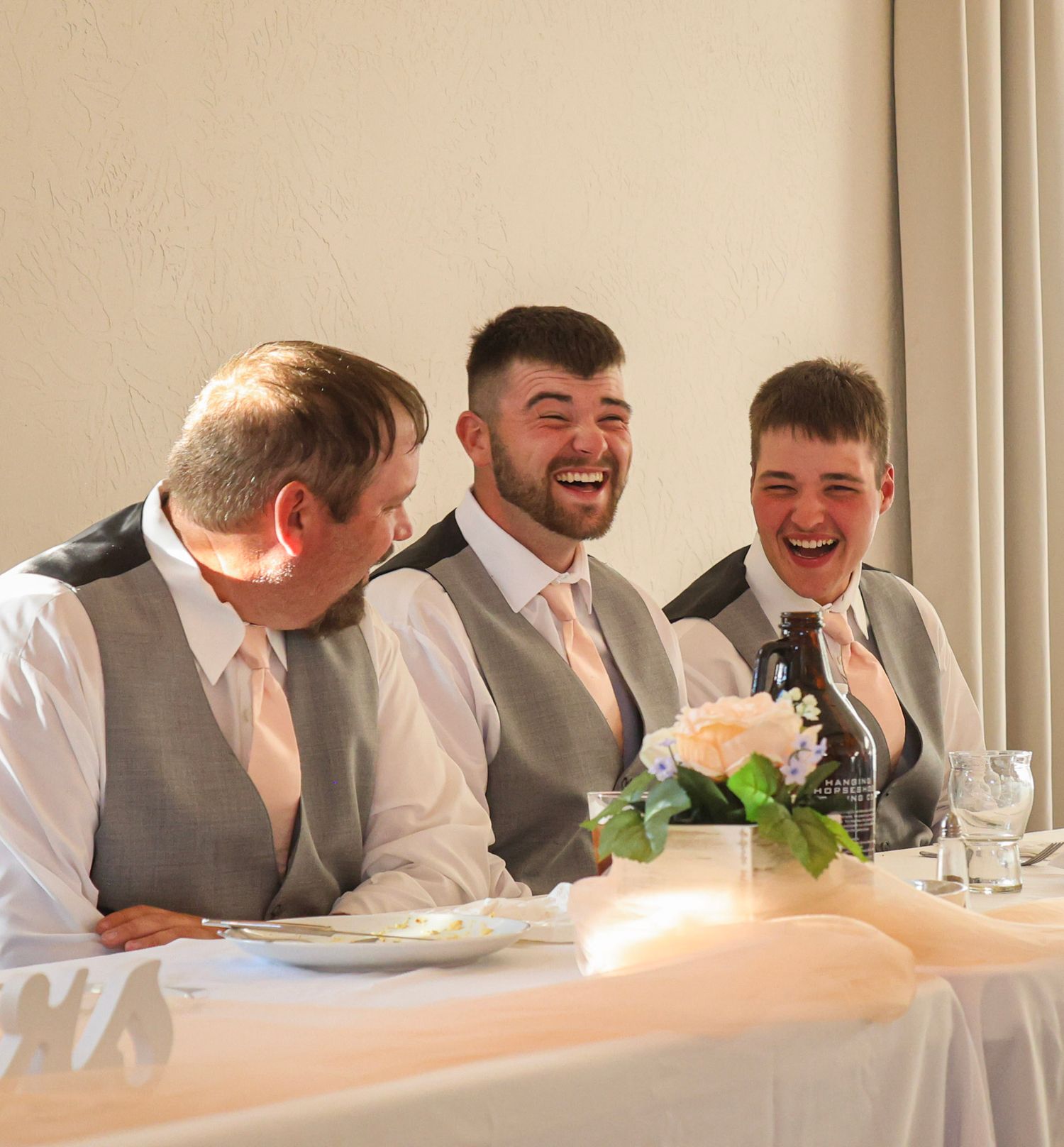 Three men in grey wedding attire laughing joyfully at the reception head table.