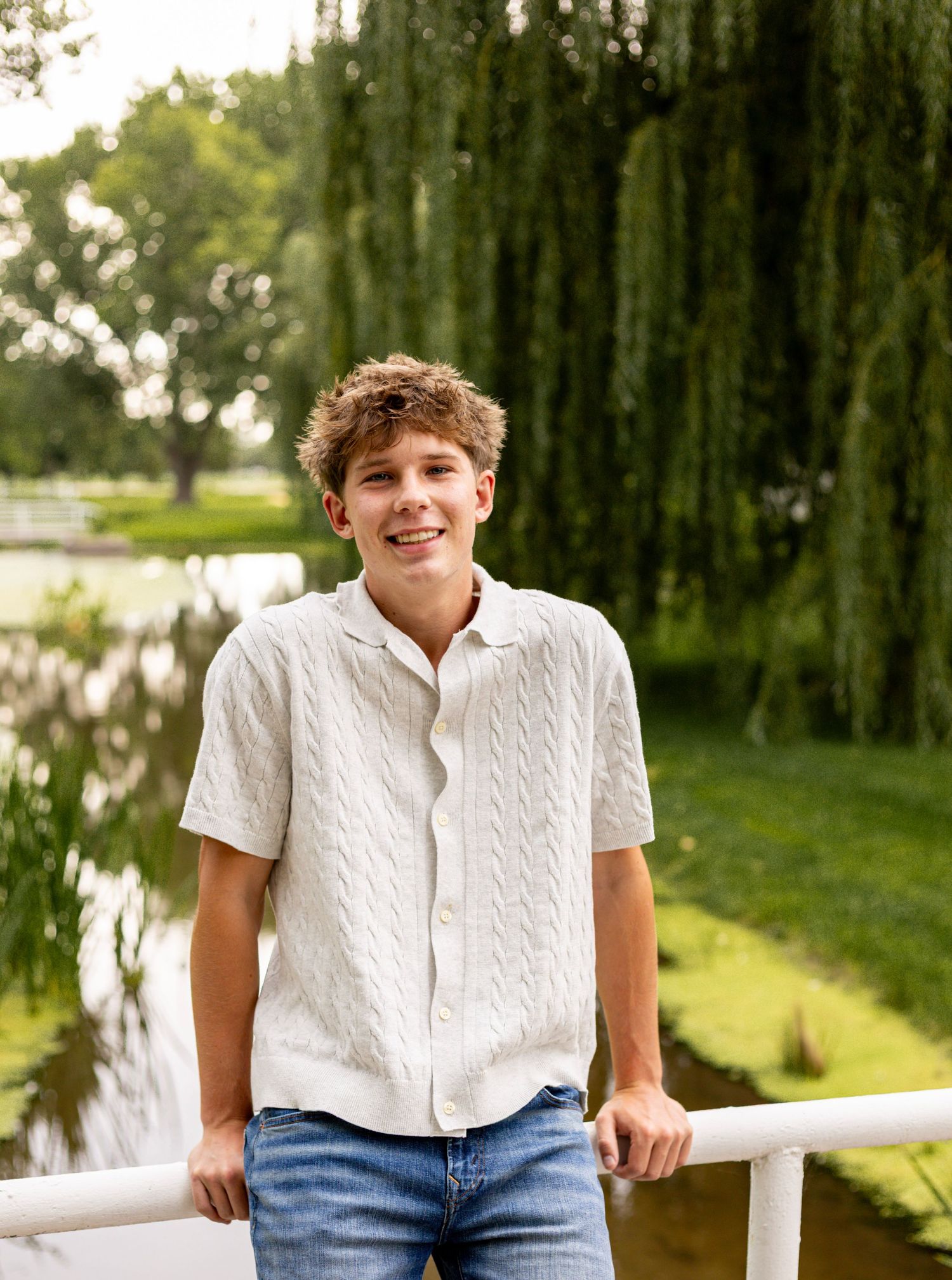 Smiling teenage boy in white knit shirt leaning on a railing near a pond with a weeping willow tree.