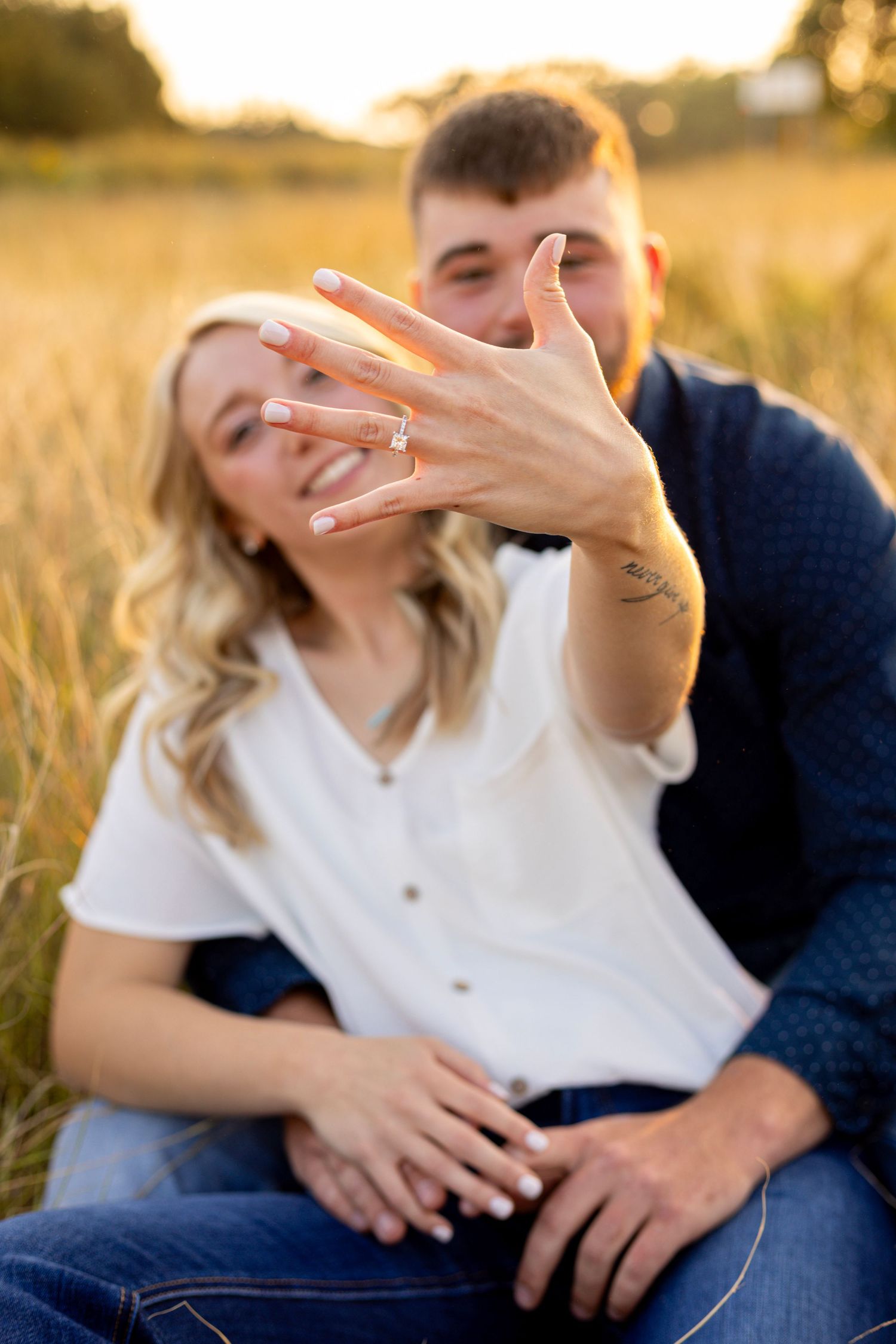A smiling woman proudly displays her diamond engagement ring while sitting with her fiance in a field.