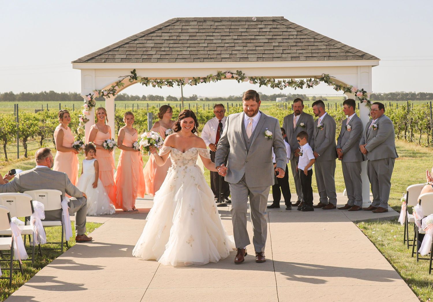 Newlyweds walk hand-in-hand down the aisle after their outdoor vineyard wedding ceremony with bridal party.