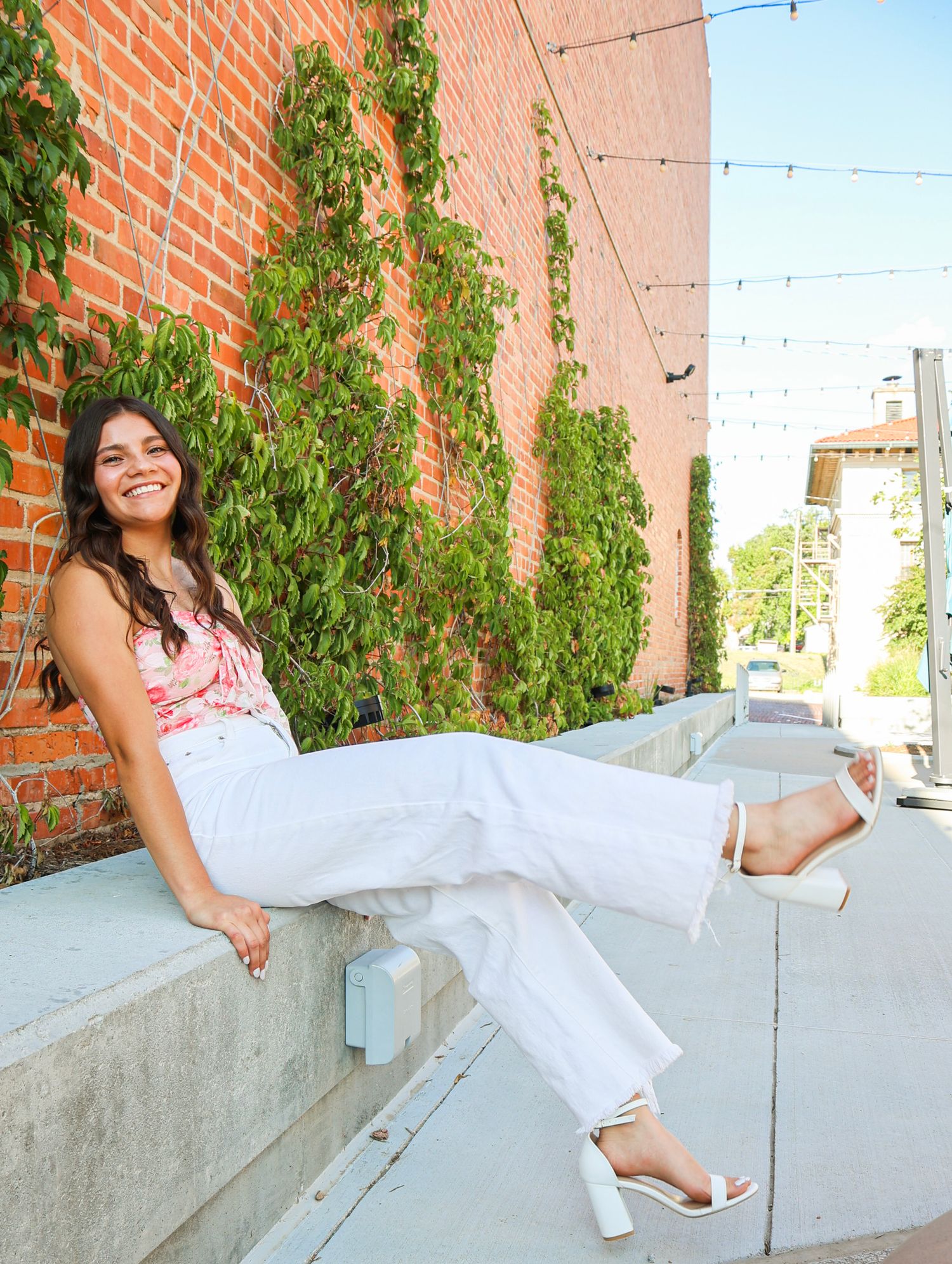 Smiling young woman in pink floral top and white pants sitting on a ledge by a brick wall with ivy.