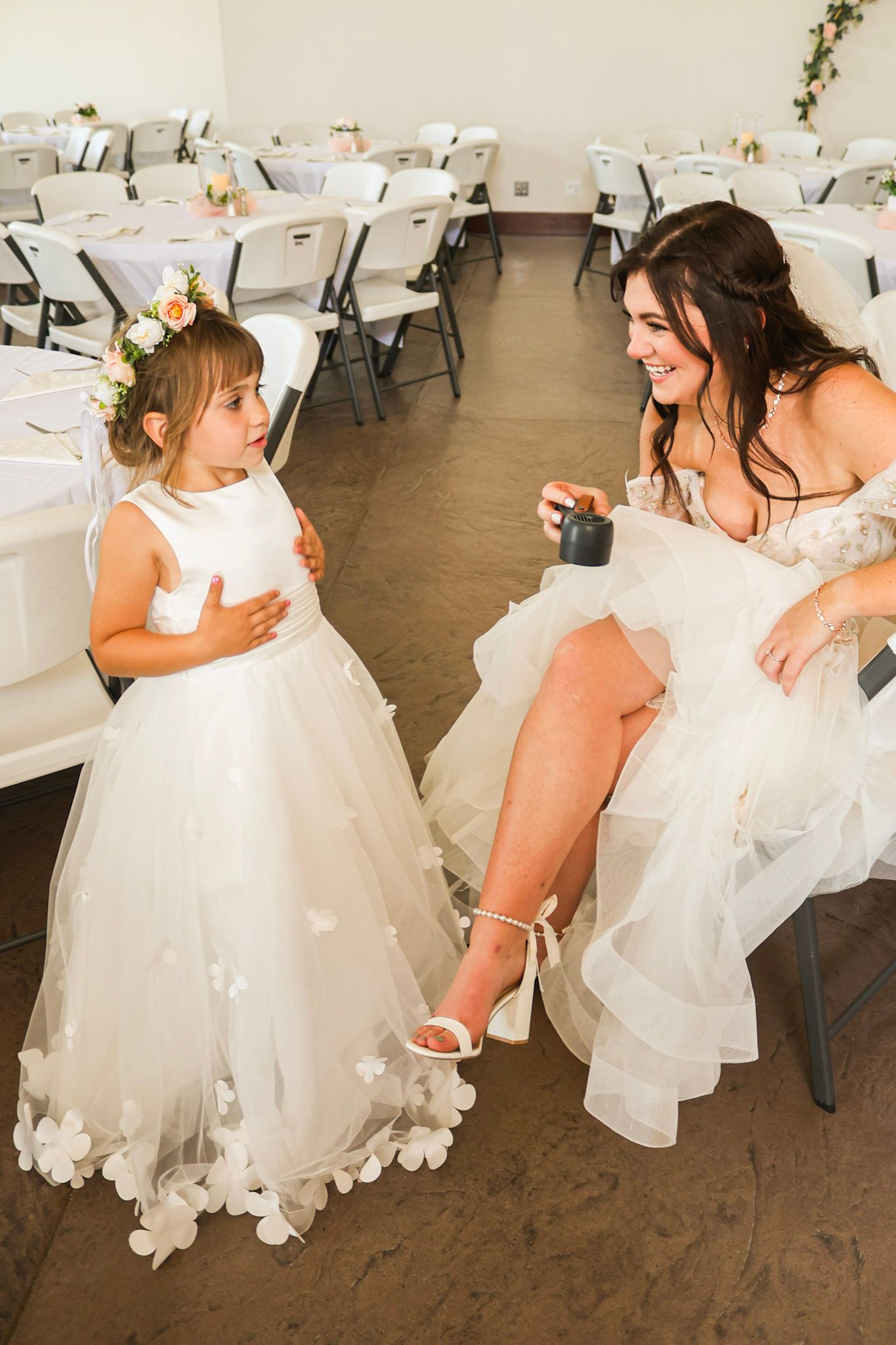 A bride in a white gown smiles and chats with a flower girl in a white dress before the reception.