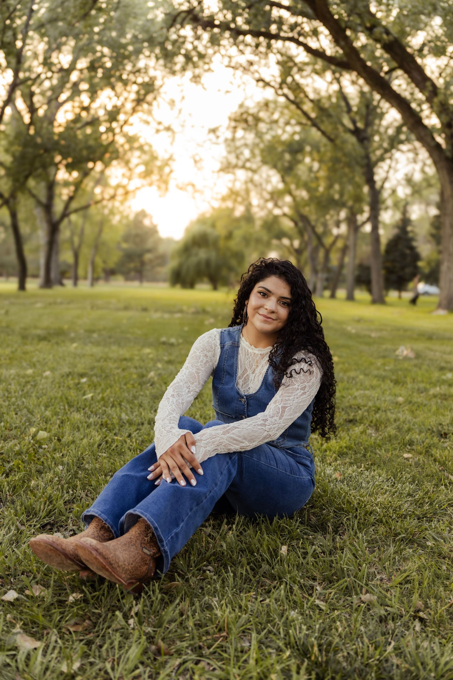Young woman with curly hair sits on green grass in a sunny park wearing denim overalls and brown boots.