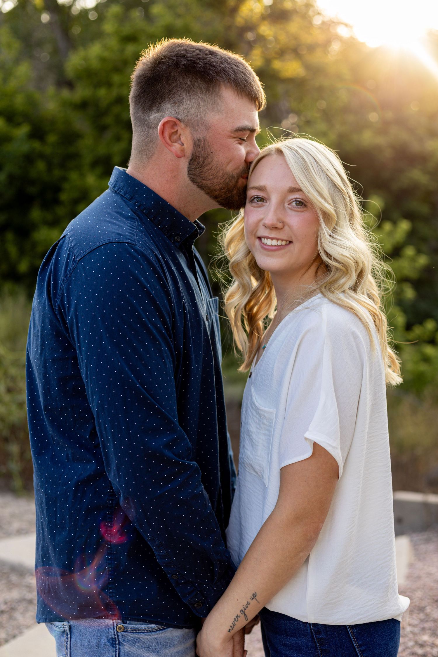 A couple shares a tender moment at golden hour, man kissing womans forehead as she smiles warmly outdoors.