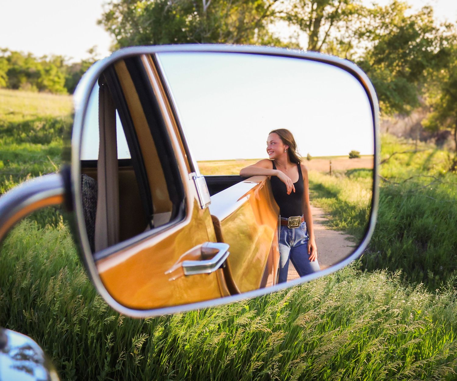 Young woman leaning on a vintage truck reflected in a side mirror on a scenic country road.