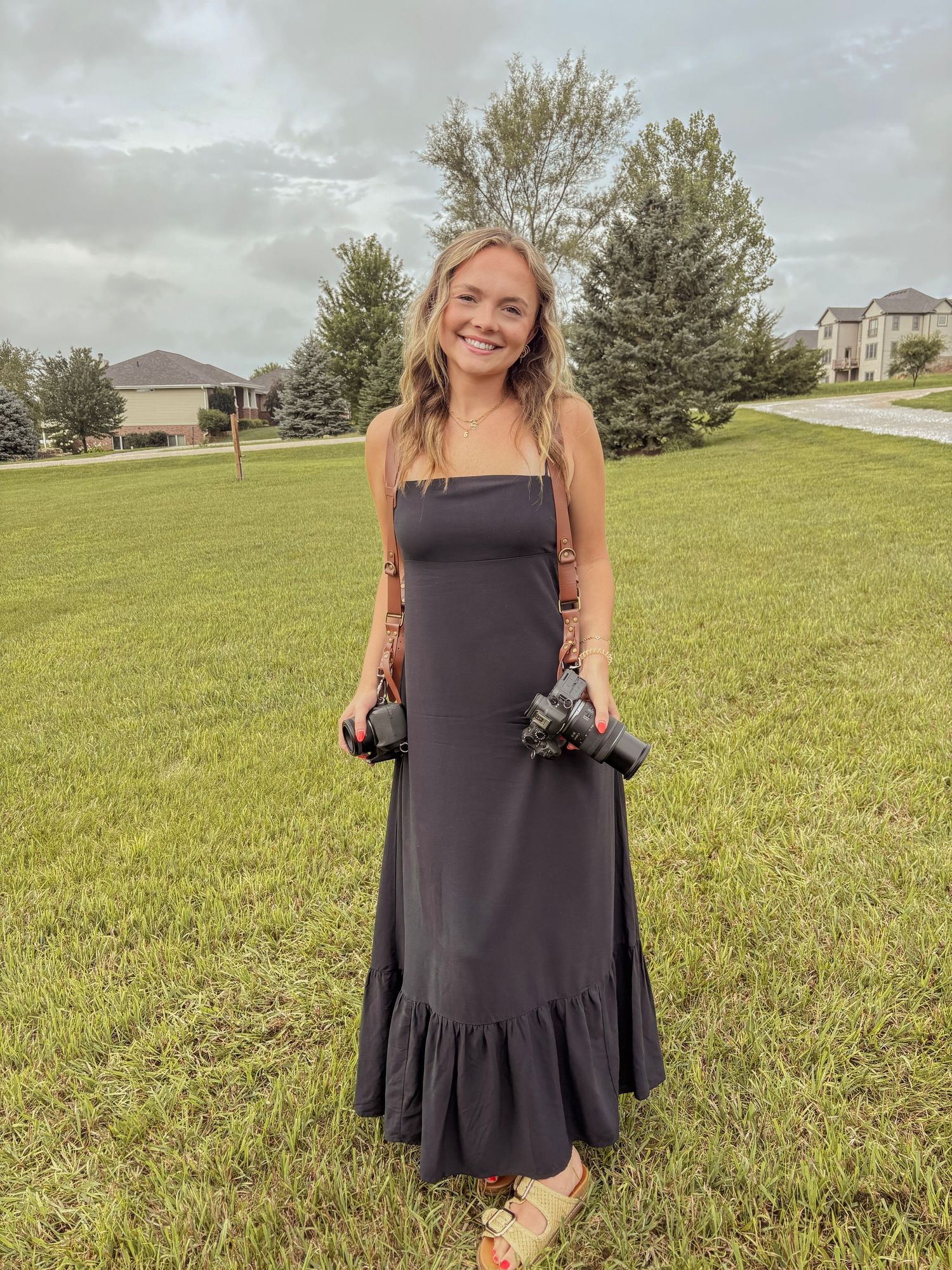 Smiling blonde woman in black maxi dress holding cameras, standing on green lawn with houses and trees in background.
