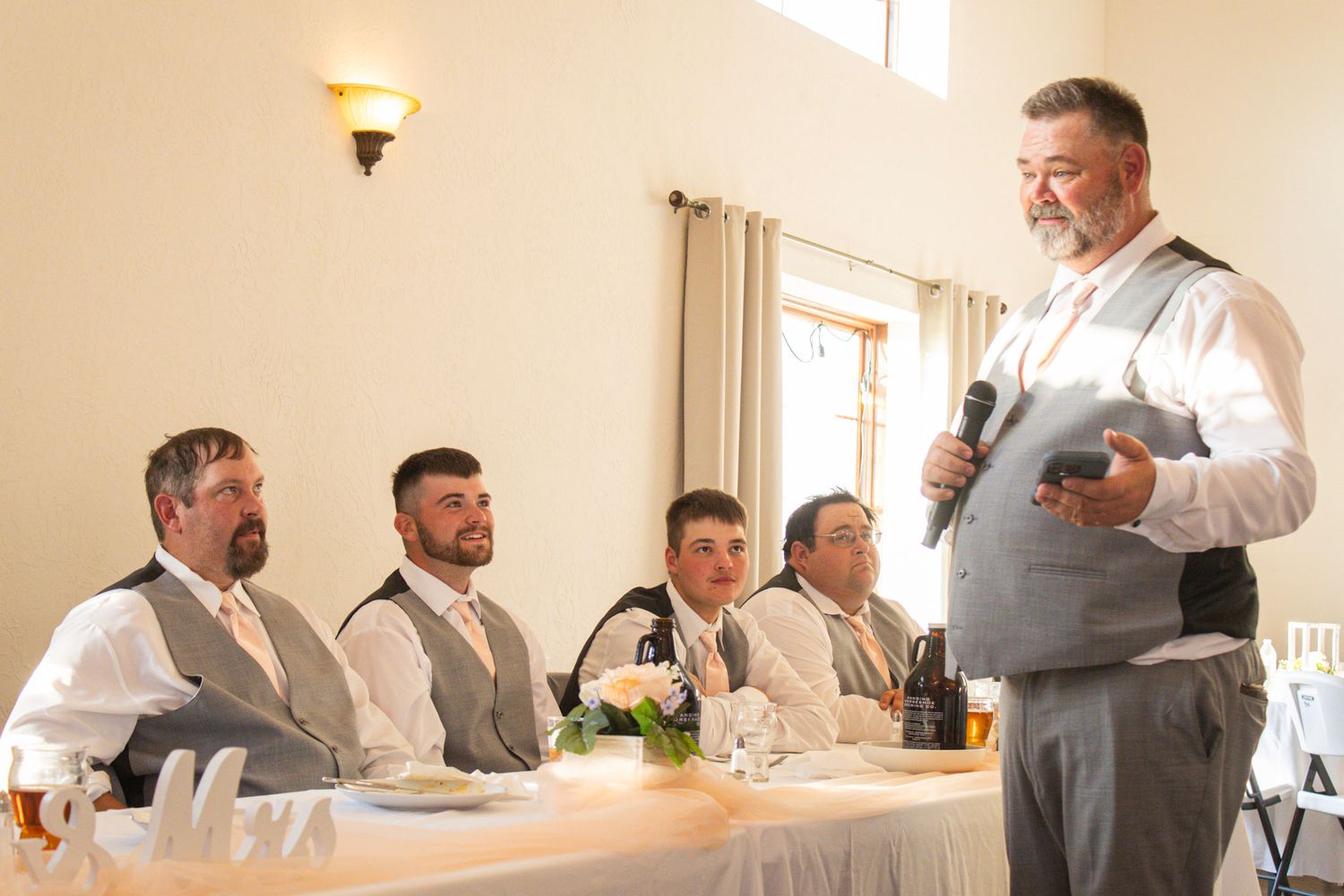 A bearded man in a gray vest gives a wedding toast while groomsmen listen at a decorated head table.