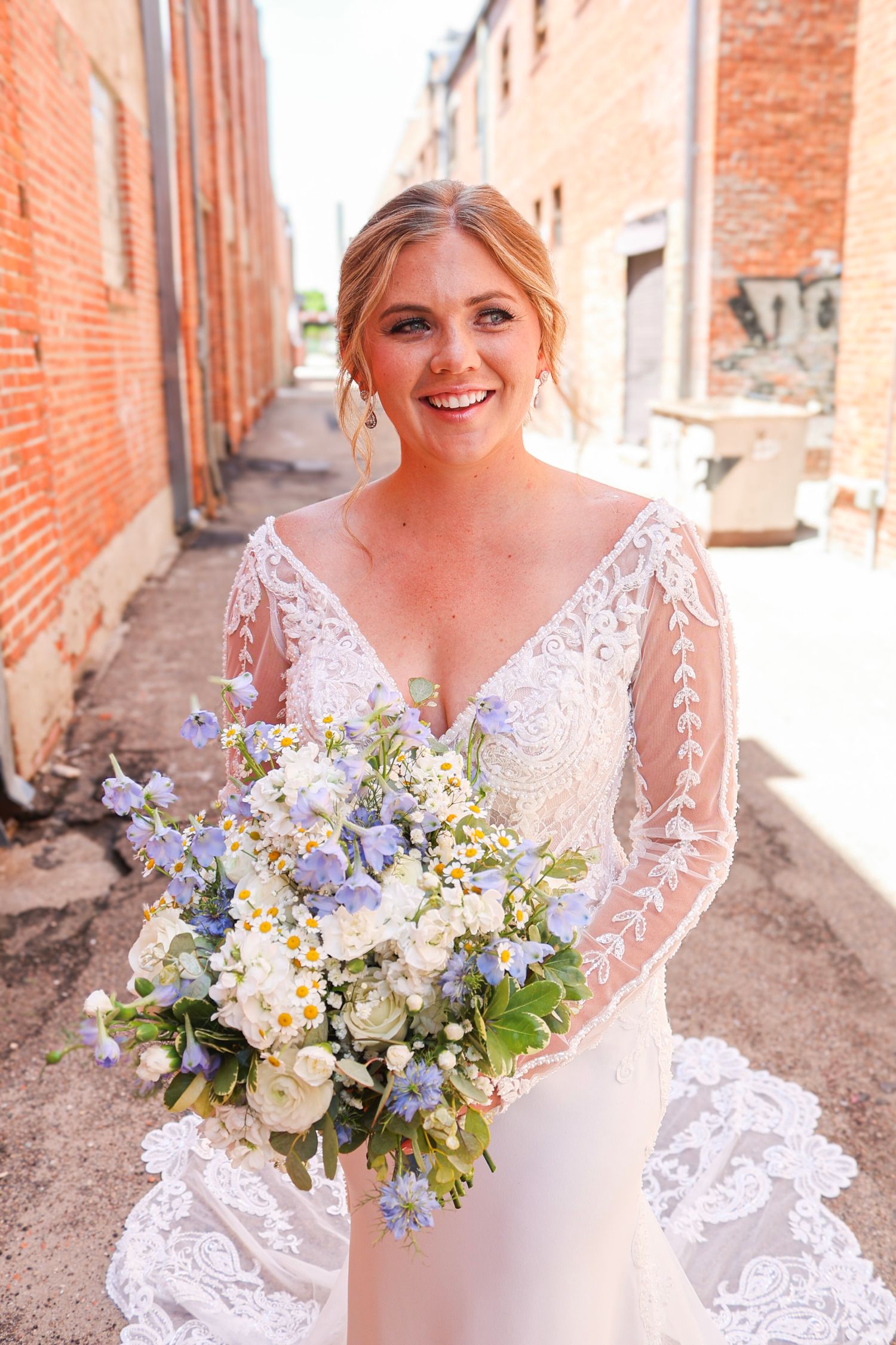 Smiling blonde bride in long-sleeve lace wedding gown holds blue and white wildflower bouquet outdoors.