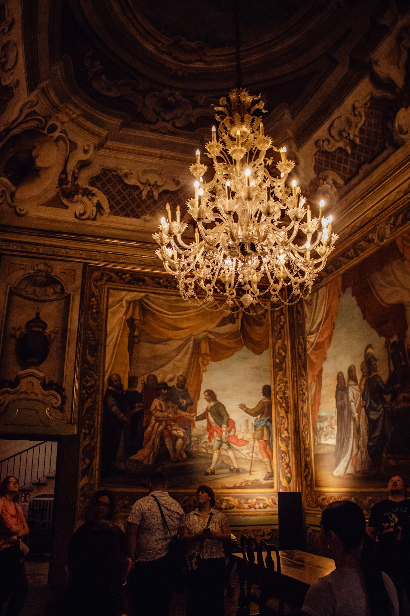 Ornate baroque interior with grand crystal chandelier, elaborate ceiling frescoes, and large classical paintings on gilded walls.