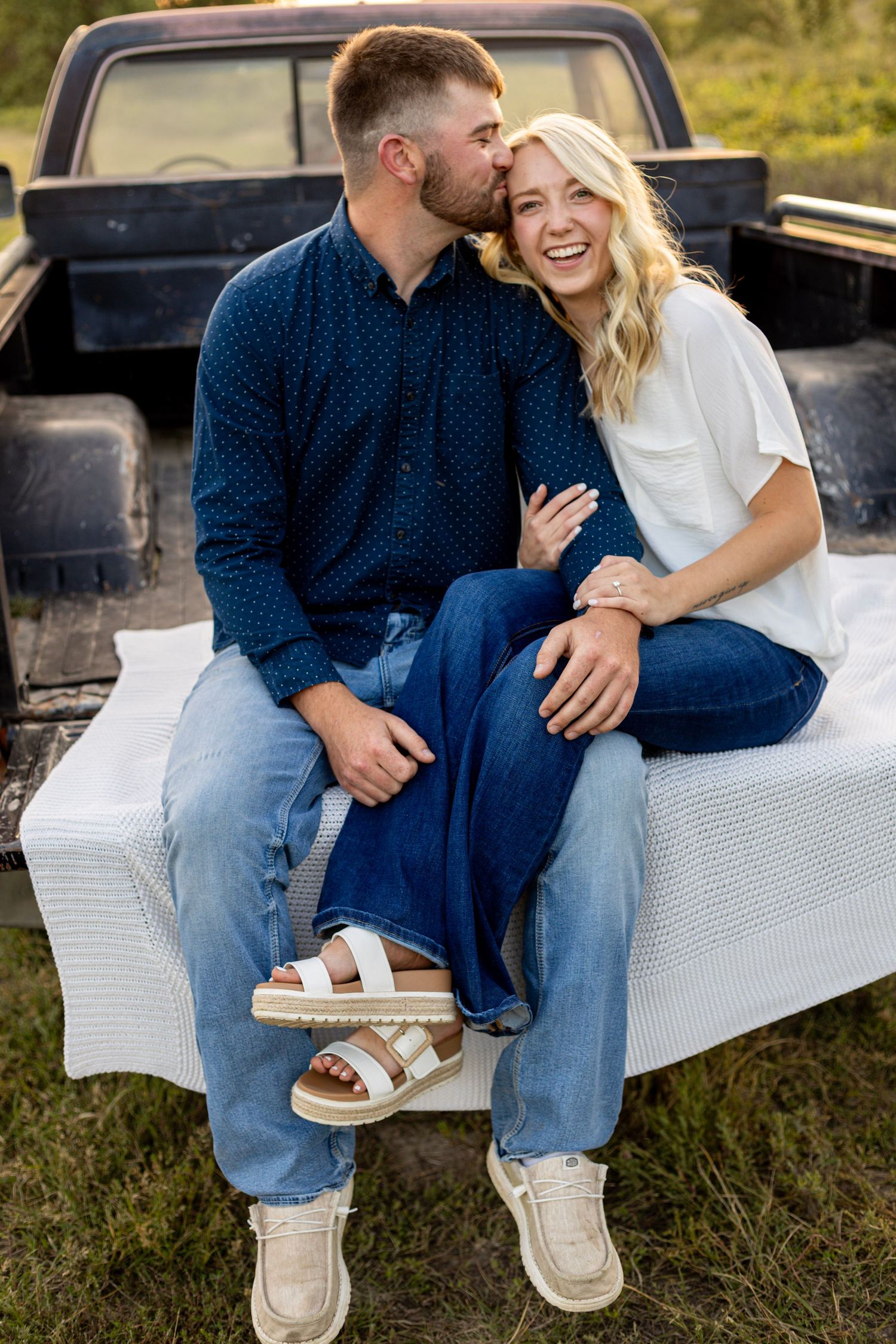 Laughing couple sitting on vintage truck tailgate during outdoor engagement photo session.