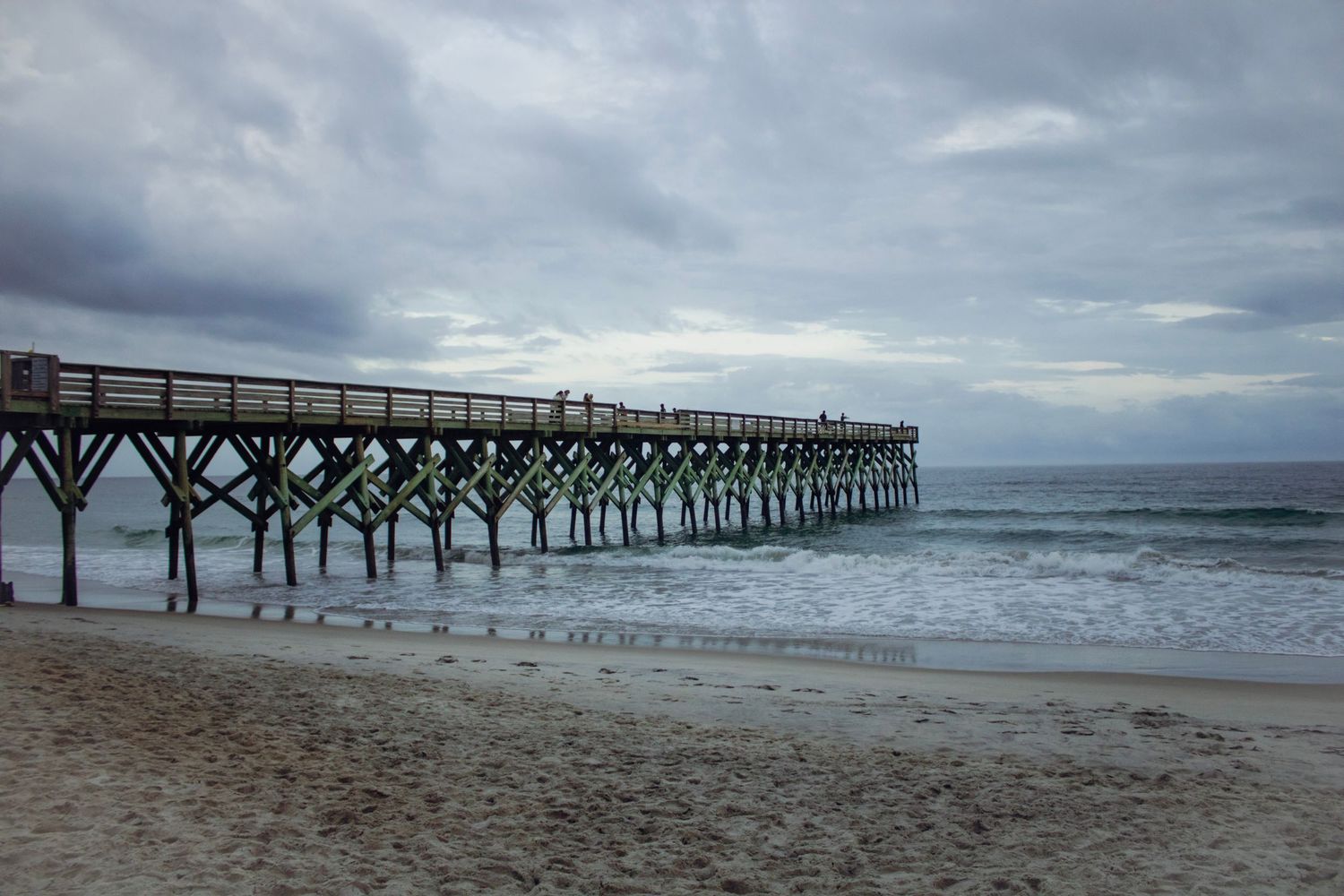 Wooden beach pier extending into the ocean under dramatic cloudy skies on a sandy shoreline.