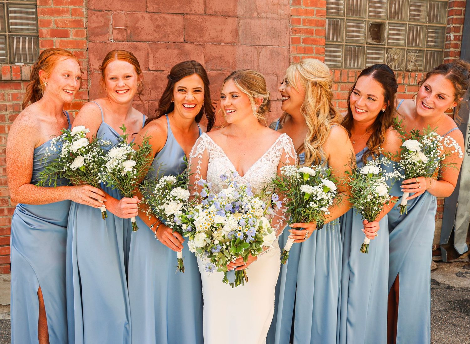 Bride surrounded by five bridesmaids in blue dresses laughing together against a brick wall backdrop.