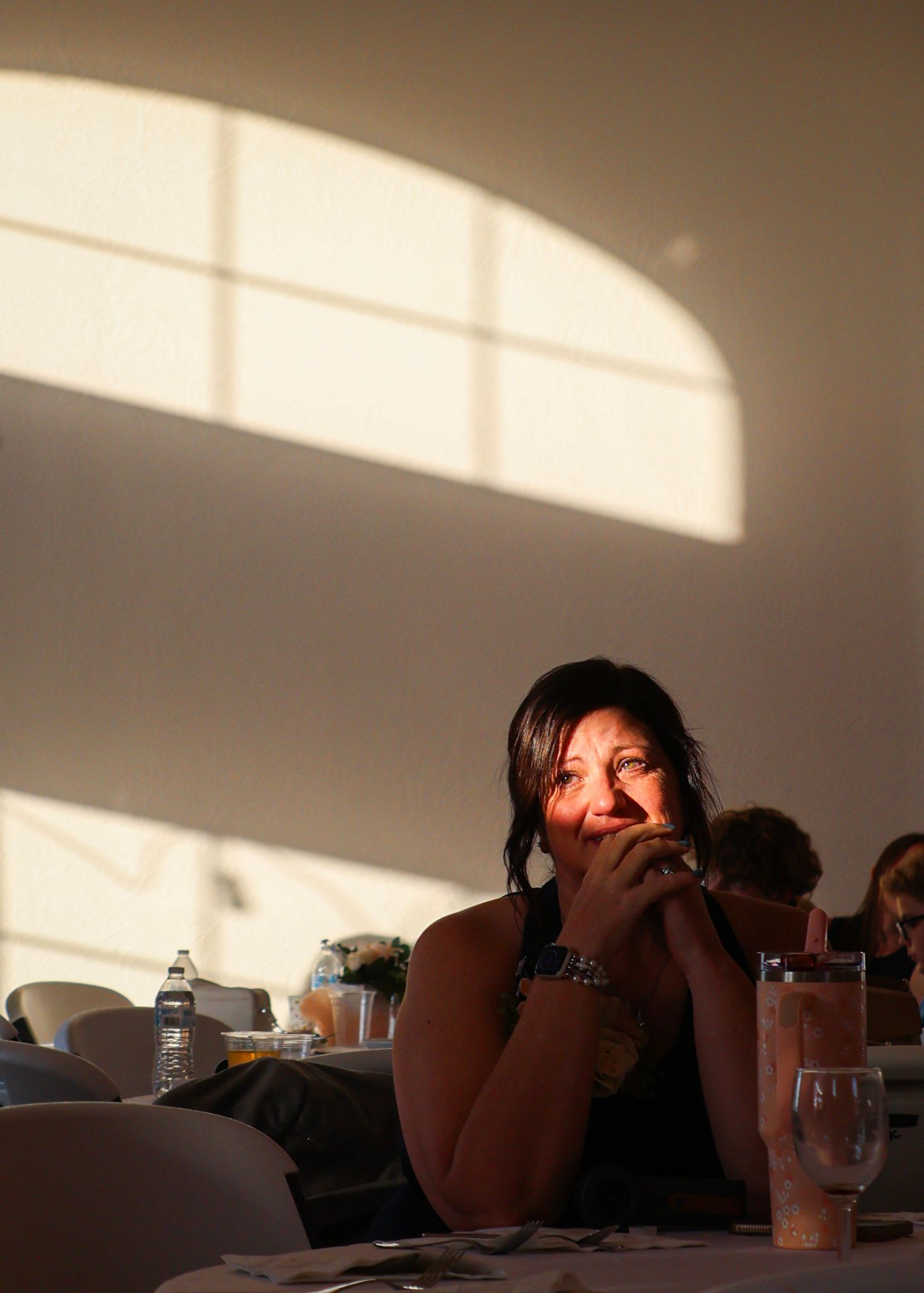 A woman smiles warmly while seated at a wedding reception table, bathed in dramatic natural light.