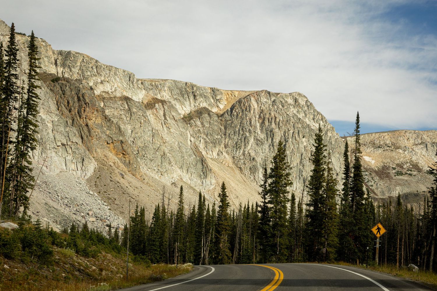 Winding mountain road flanked by evergreen trees with dramatic rocky cliff face under cloudy sky.