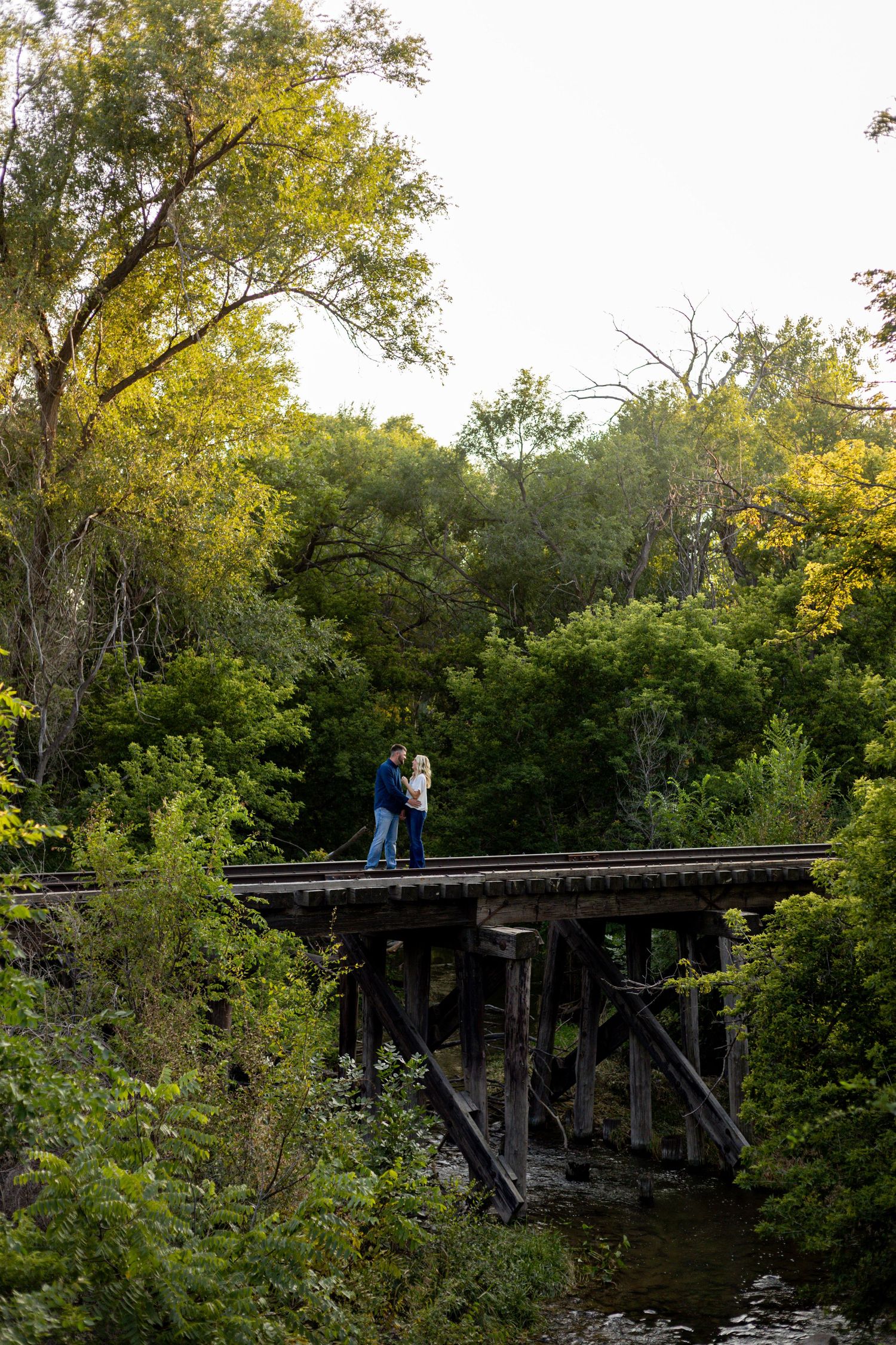 Engaged couple embracing on a rustic wooden trestle bridge surrounded by lush green autumn trees.
