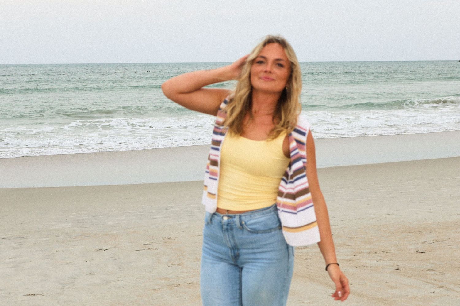 Smiling blonde woman in yellow top and jeans posing on a beach with ocean waves behind her.