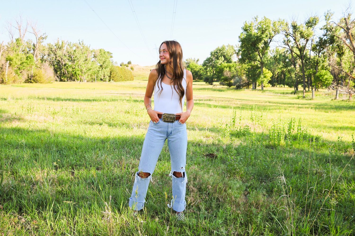 Happy young woman in white tank and ripped jeans standing in a sunlit green meadow with trees.