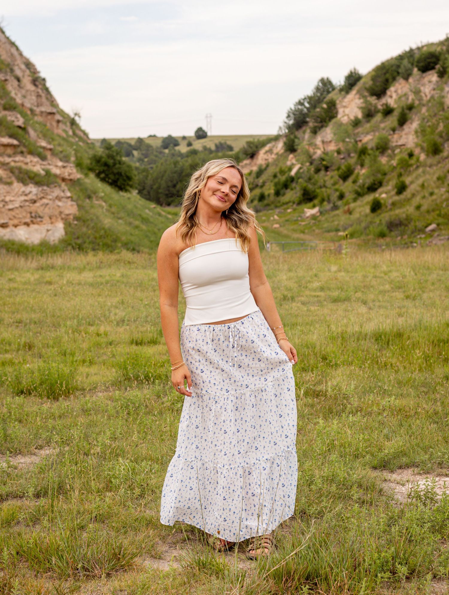 Young woman in white tube top and floral maxi skirt posing in a green meadow with rocky hills behind her.