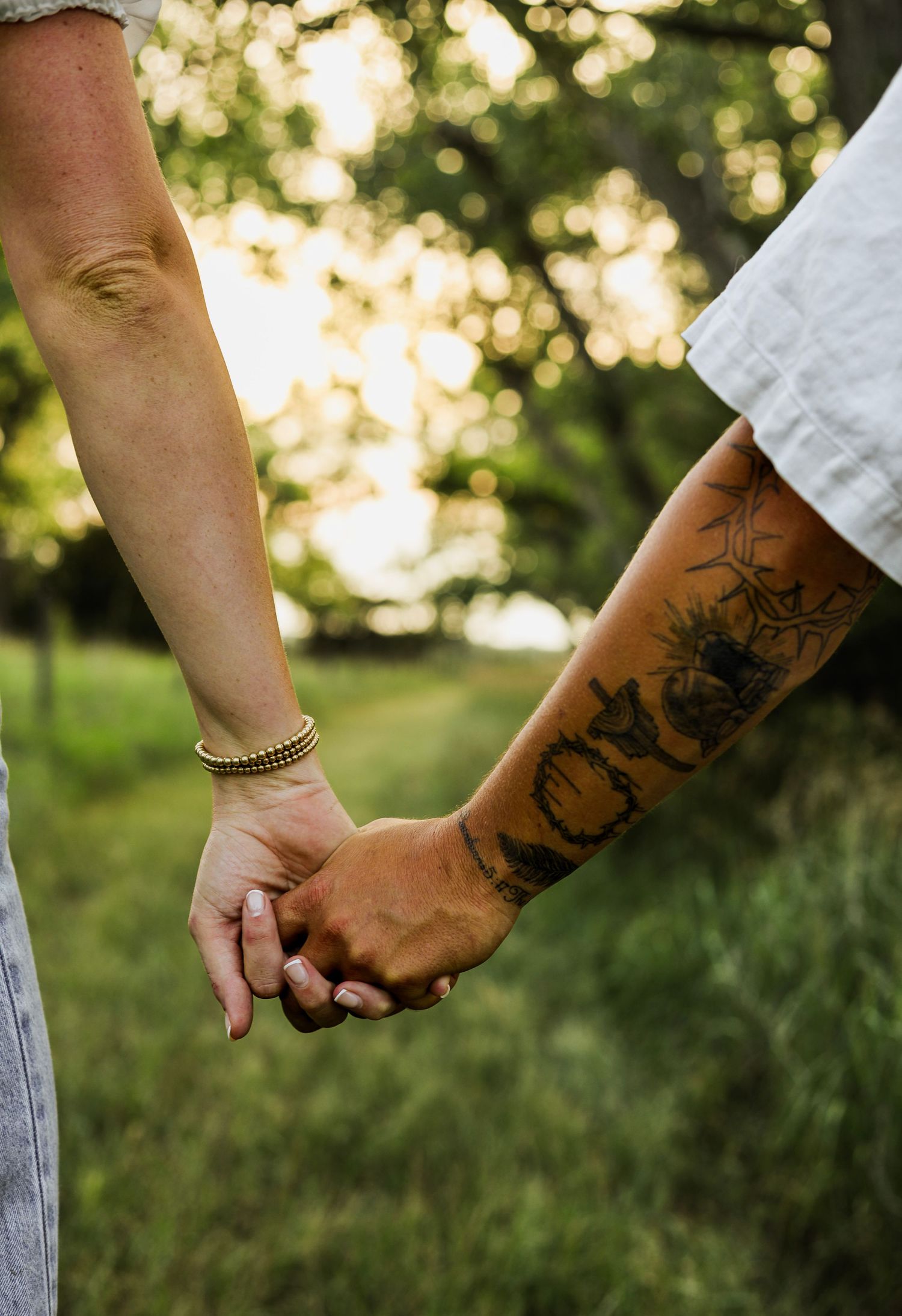 Couple holding hands outdoors, one with floral forearm tattoo and other wearing gold bracelet, green bokeh background.