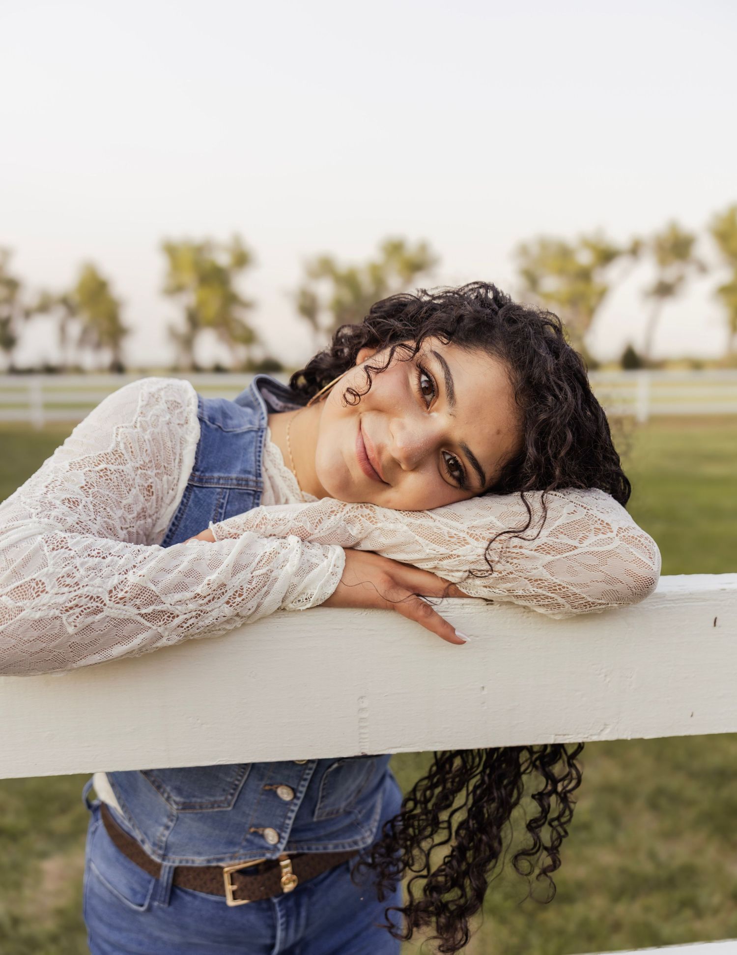Young woman with curly hair leans on white farm fence wearing denim vest and lace top outdoors.