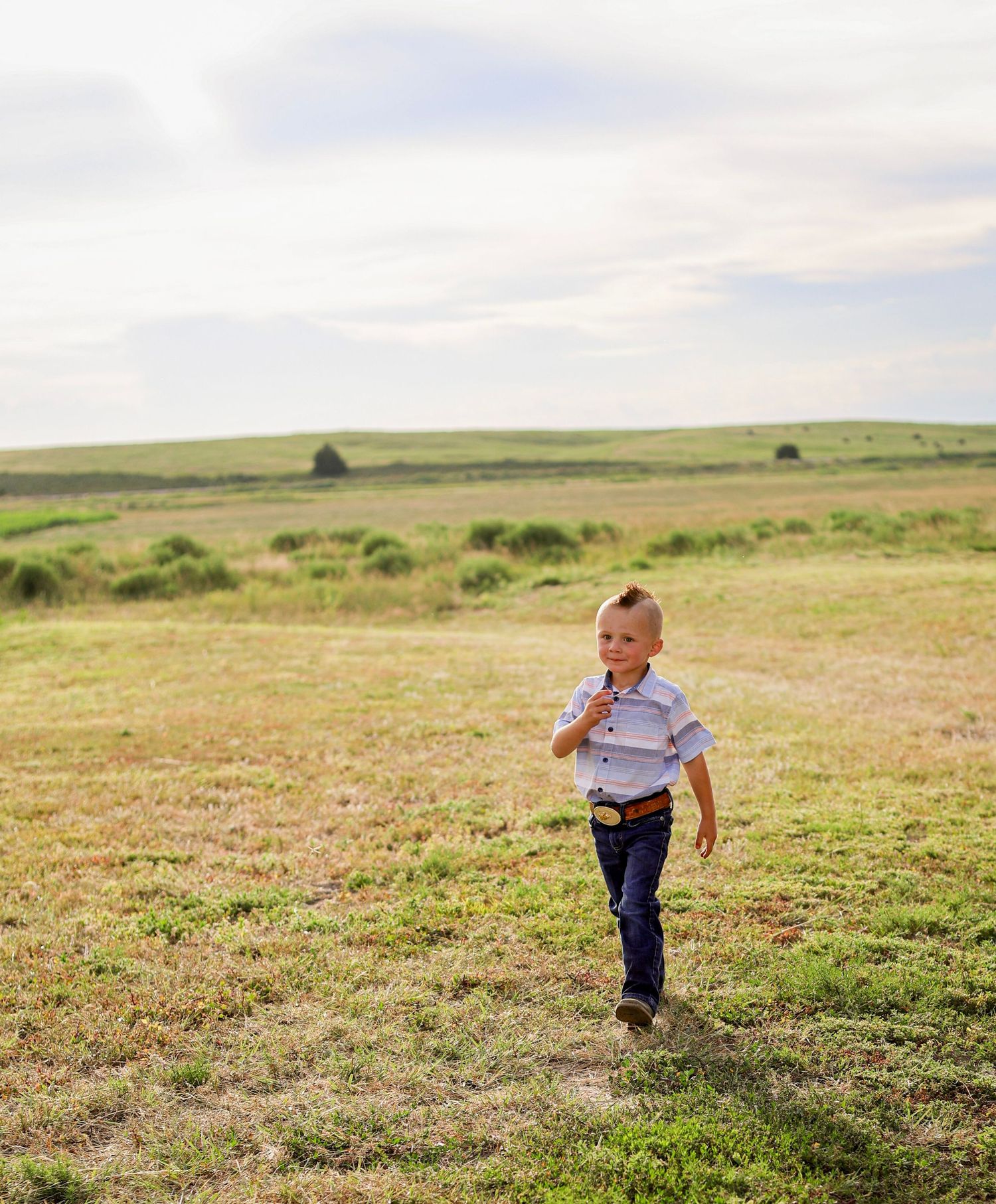 Young boy in white shirt running through open green field under cloudy sky.