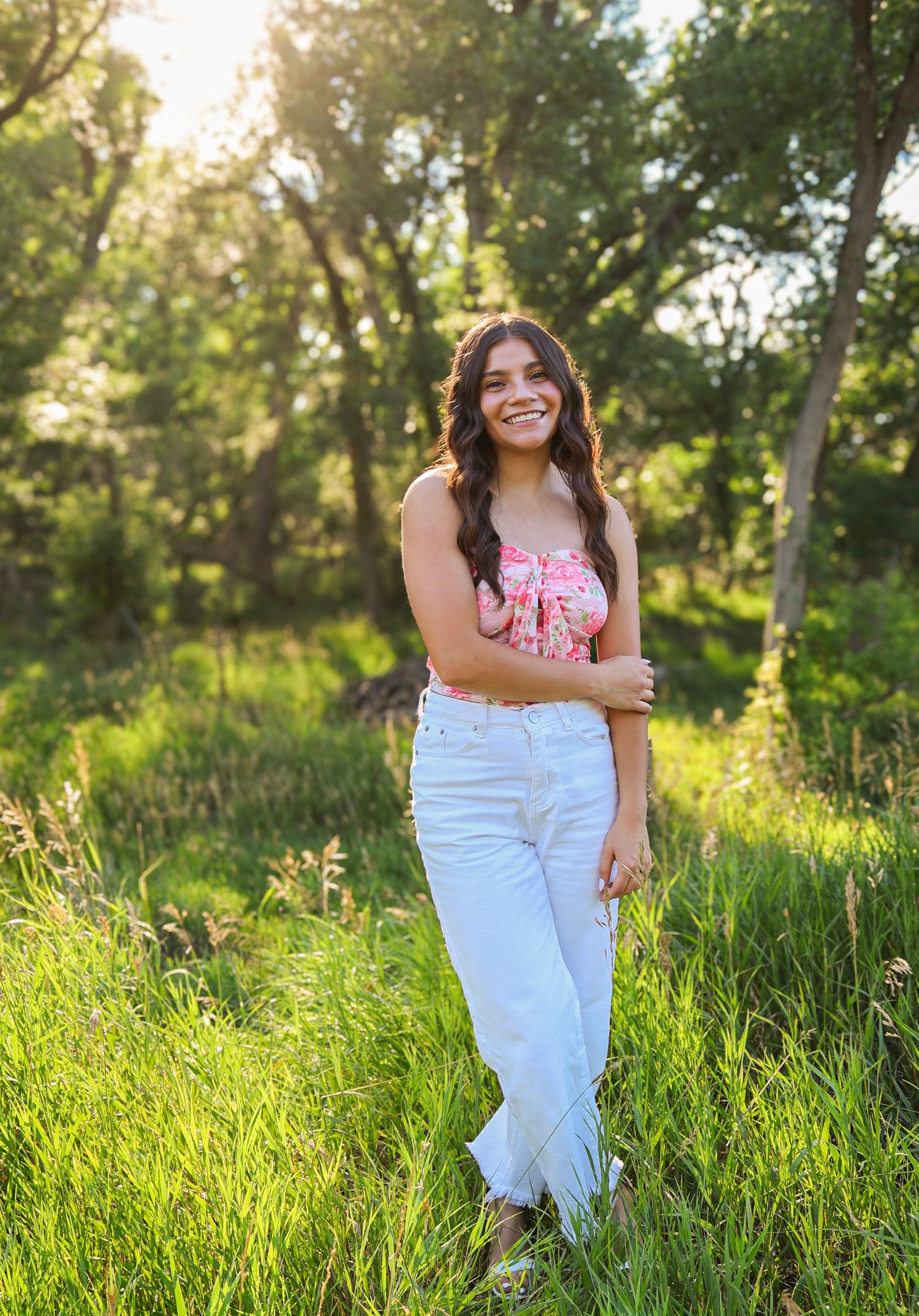 Smiling young woman in pink floral top and white jeans posing in a lush green outdoor setting.