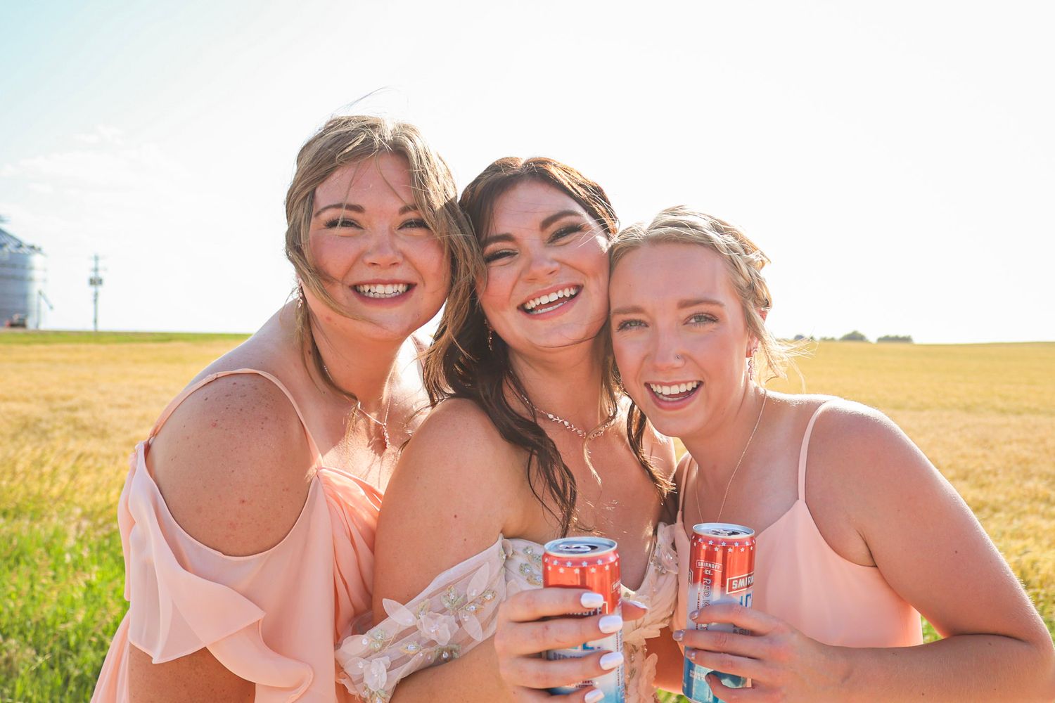 Three bridesmaids in blush dresses laughing and holding drinks in a sunny field.