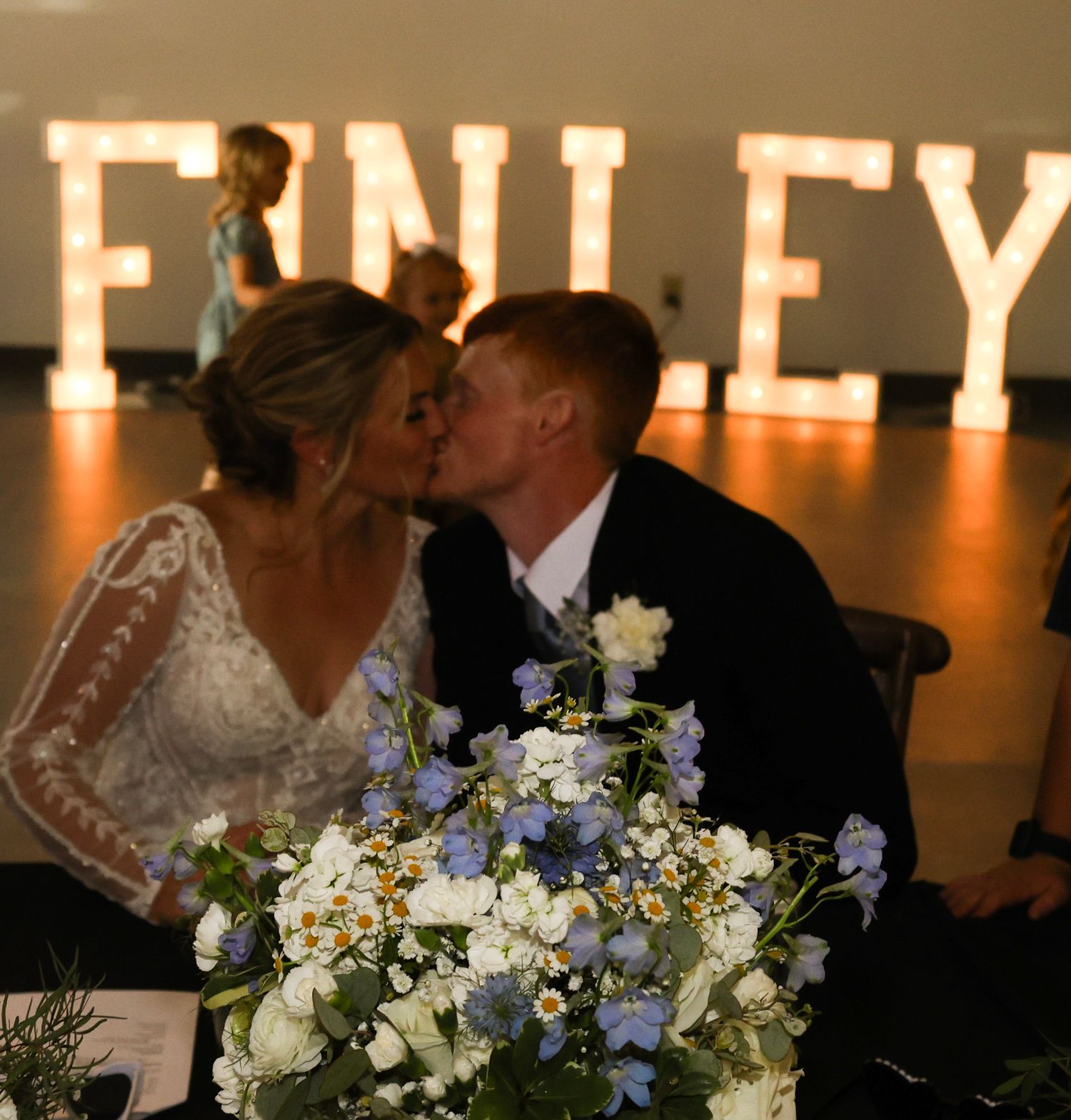 Bride and groom share a kiss at sweetheart table in front of glowing FINLEY marquee letter backdrop.