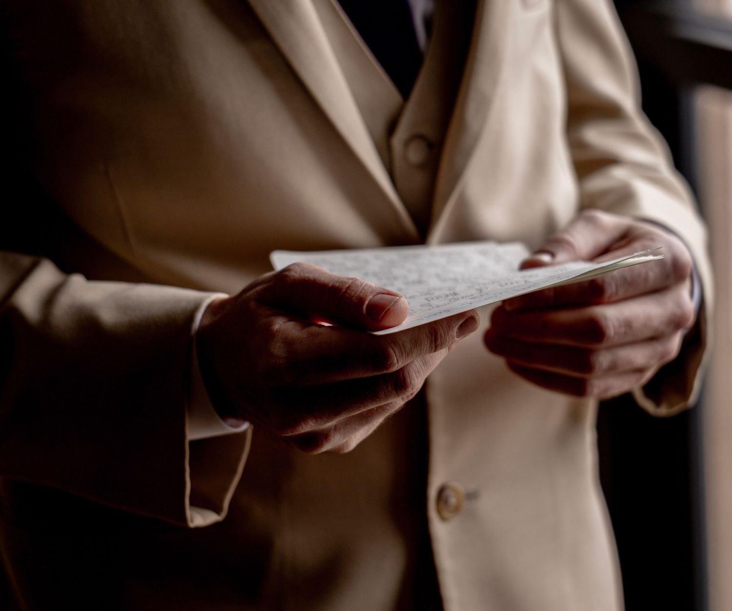 Person in beige blazer reading a handwritten letter or note in dimly lit indoor setting.