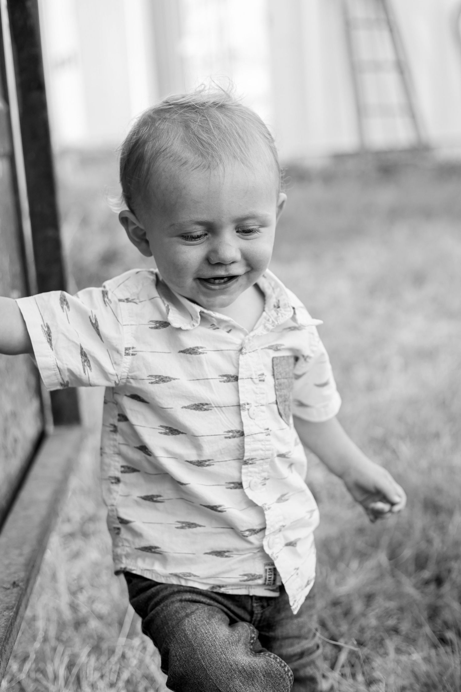Black and white photo of a happy toddler boy laughing outdoors, wearing a patterned shirt and jeans.