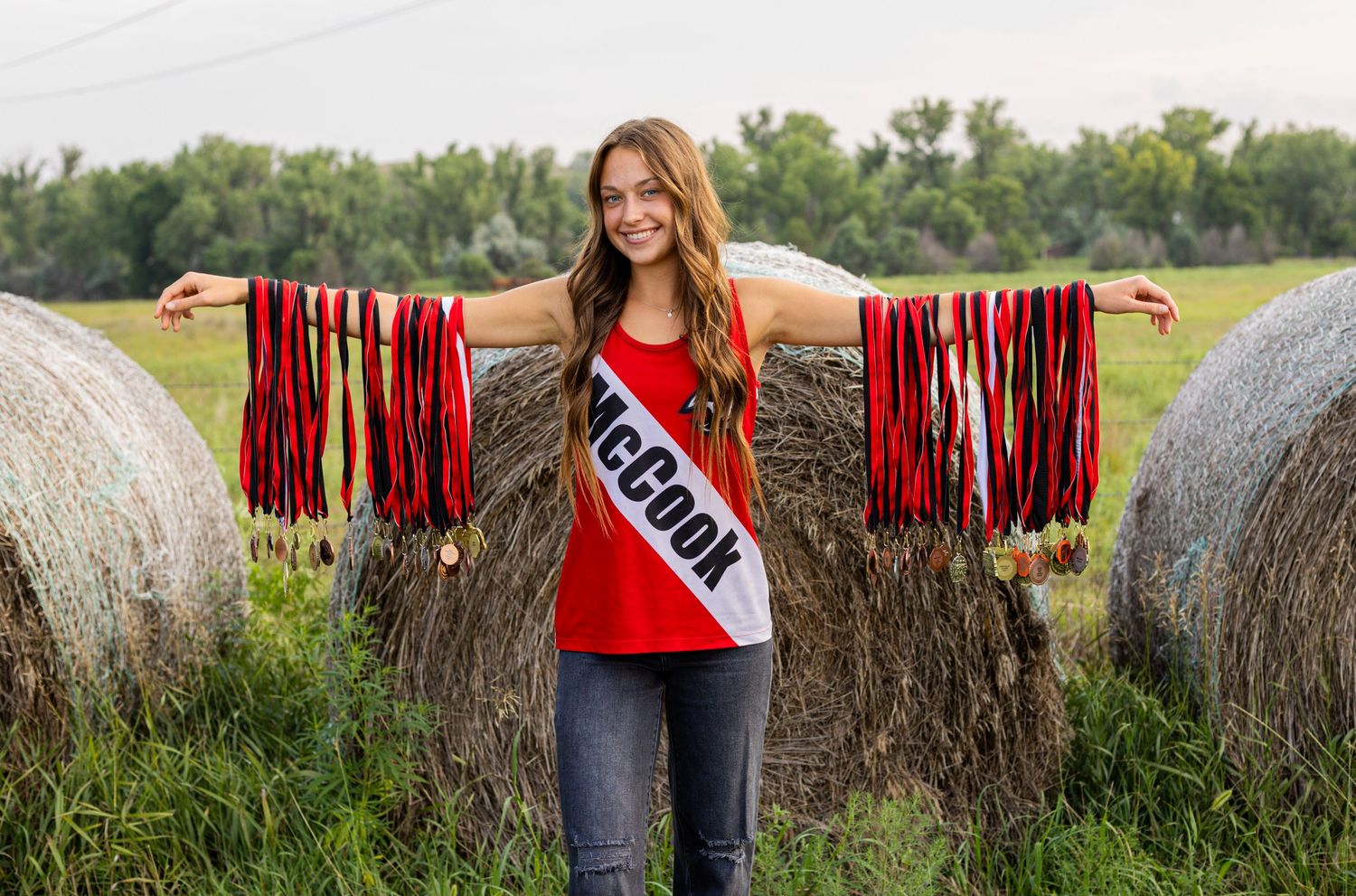Smiling young woman wearing McCook sash displaying long red medal ribbons beside hay bales in field.