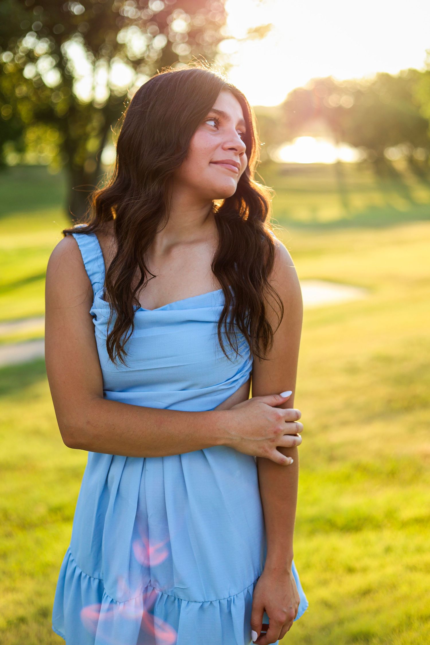 Young woman in light blue sundress standing in golden sunset light near a serene pond outdoors.