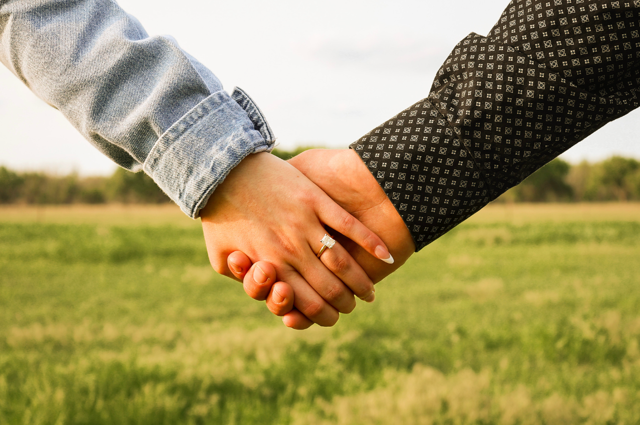 Close-up of couple holding hands showing engagement ring in green field, wearing denim and black dress shirts.