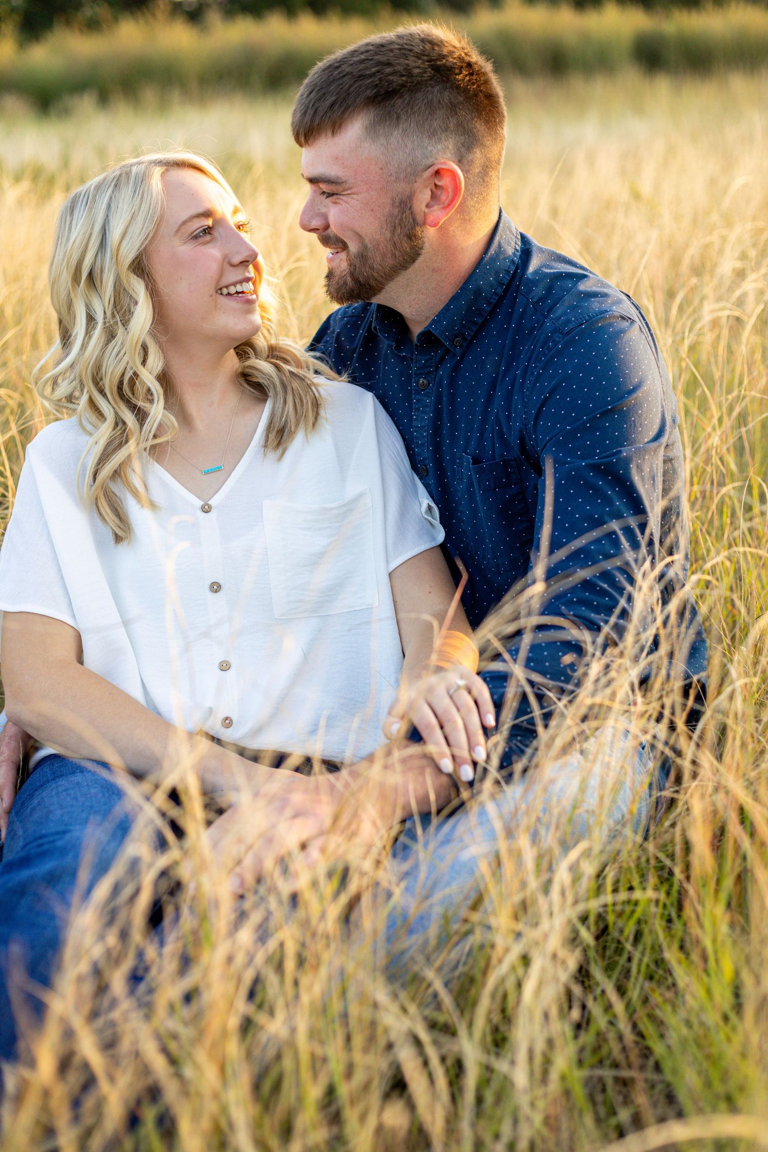 A smiling engaged couple sits together in tall golden grass, gazing lovingly at each other.