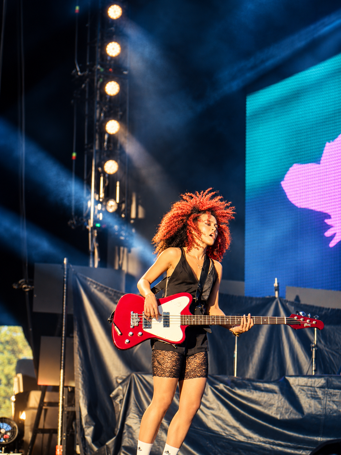 Female guitarist with curly red hair performs on stage with a red electric guitar under bright stage lights.