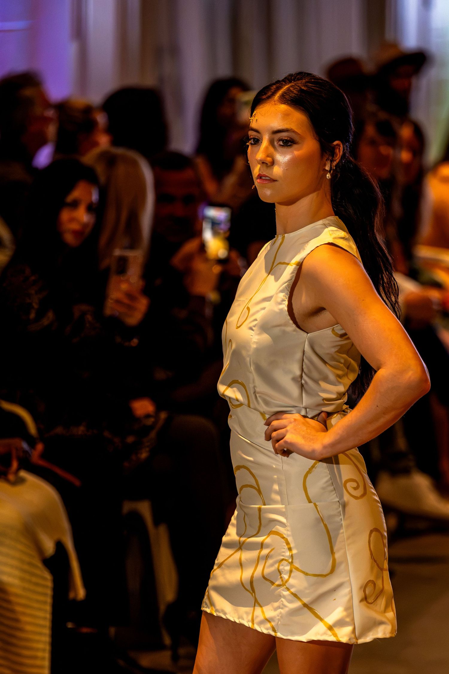 Model wearing a white and gold mini dress on the Charlotte Fashion Week runway, with guests photographing the show.