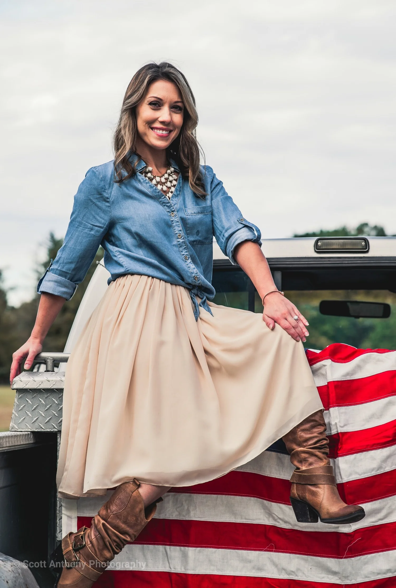 Woman in denim shirt, cream skirt, and brown boots poses on truck tailgate with American flag backdrop.