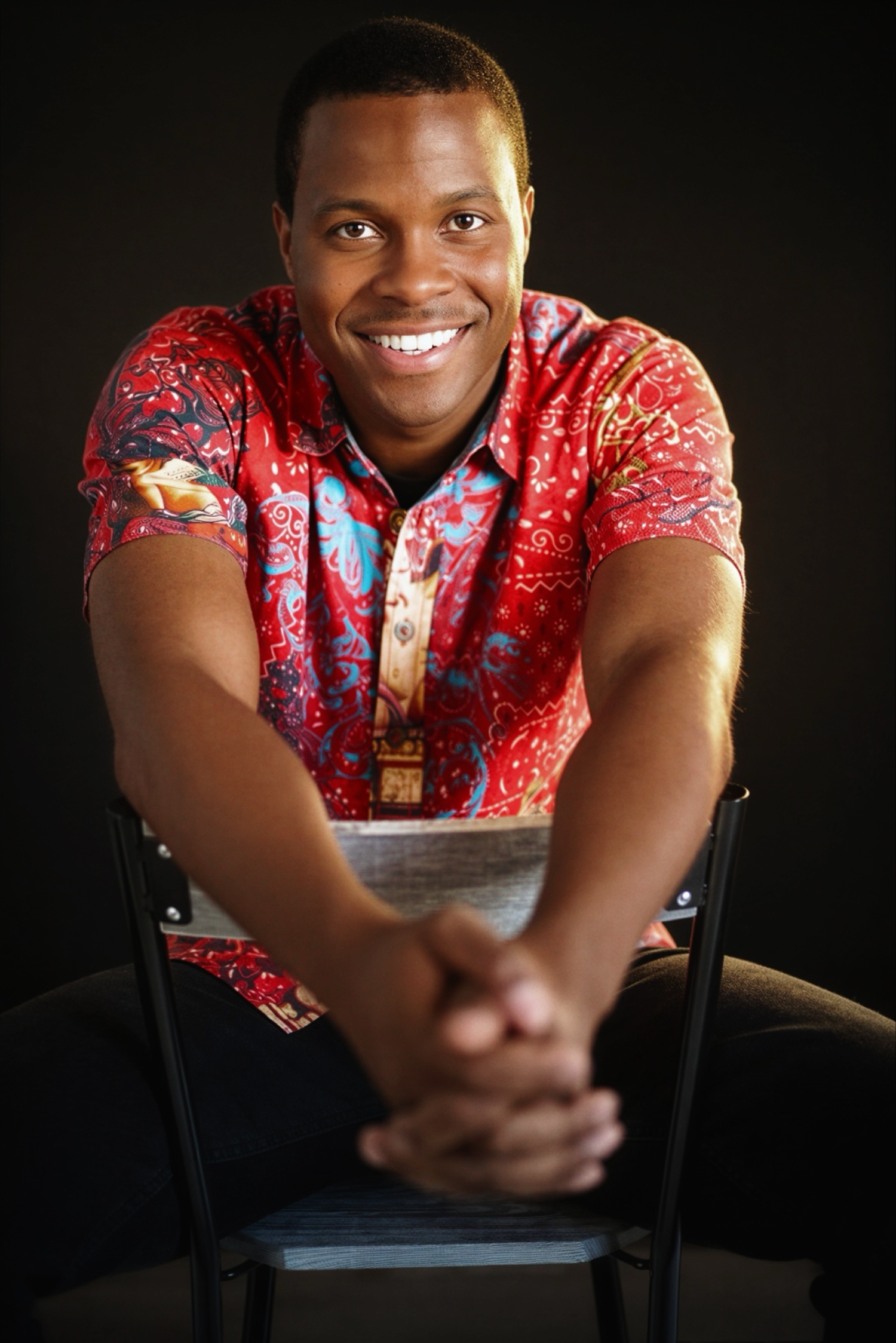 Smiling man in colorful red floral shirt sitting on chair with hands clasped, posed against dark background.