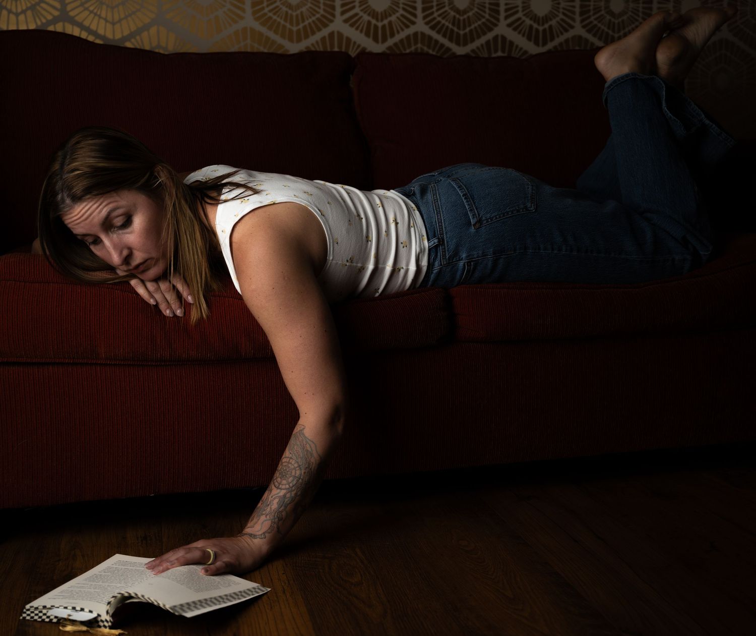 Woman lying on red couch reaching down to read a book on hardwood floor in dimly lit room.