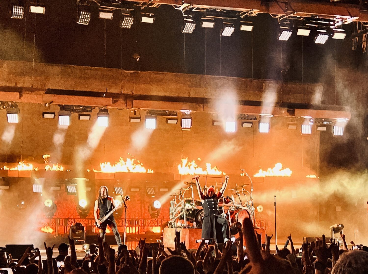 Heavy metal band performs on stage with dramatic fire effects and bright lights before a large crowd.