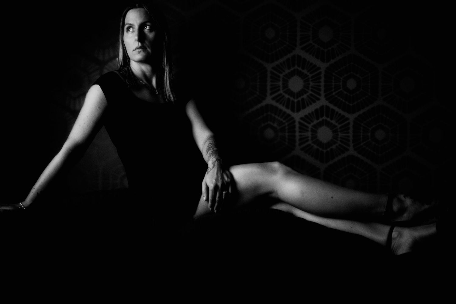 Black and white portrait of a woman in a dark studio, sitting gracefully with geometric patterns in the background.