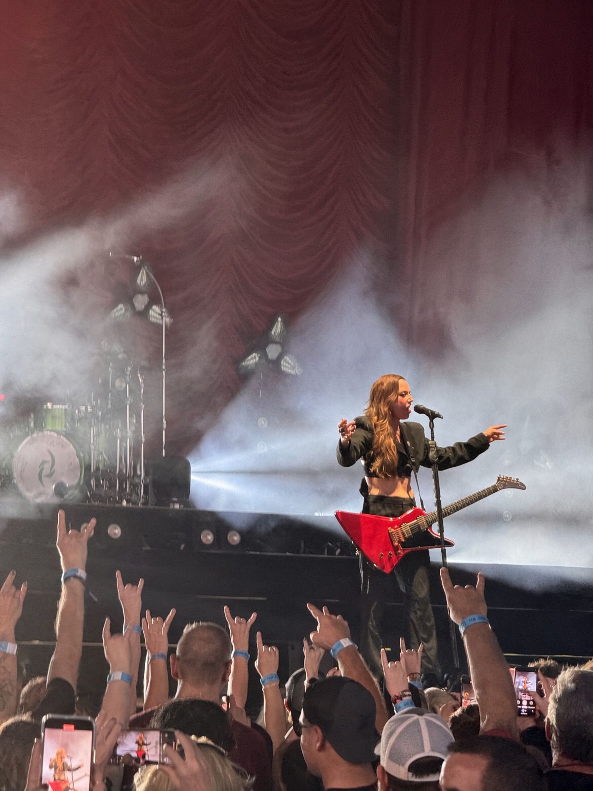 Female guitarist with red V-shaped guitar performs on smoky stage while crowd raises hands at rock concert.