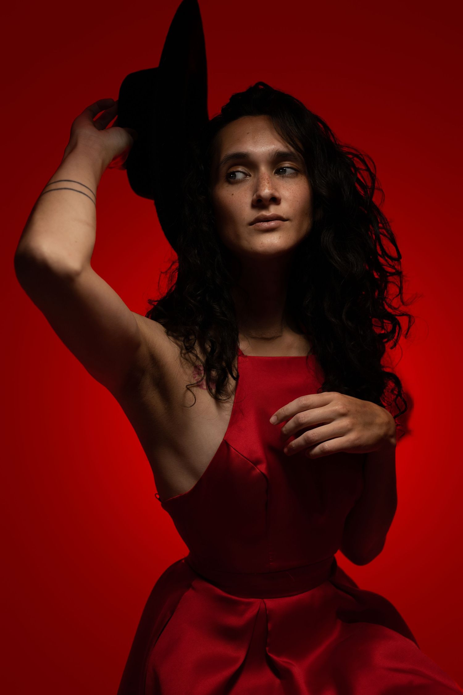 Woman in red dress holding black hat against vibrant red background in dramatic studio portrait photography.