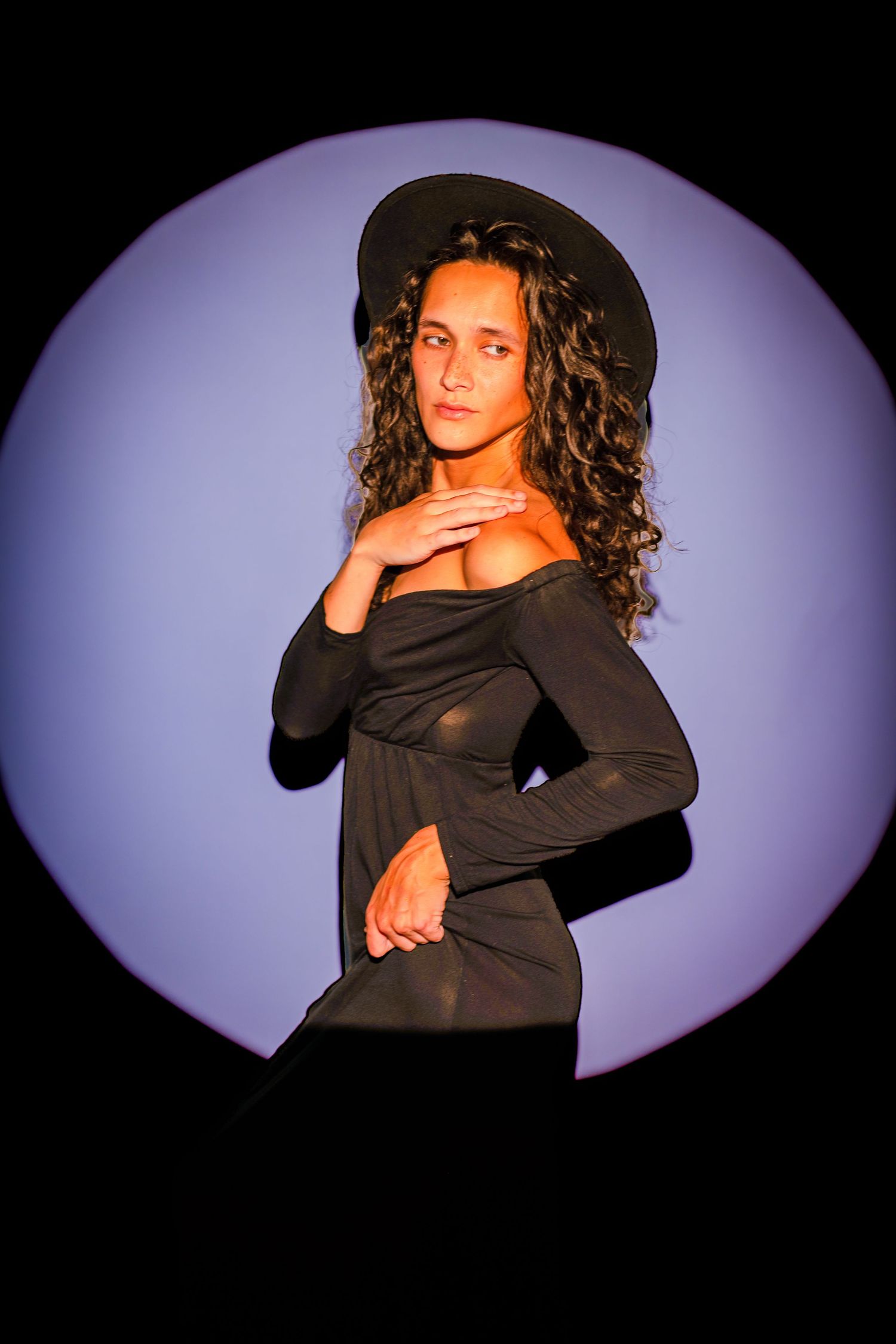 Woman in black dress and wide-brim hat poses confidently under a circular purple spotlight against dark background.