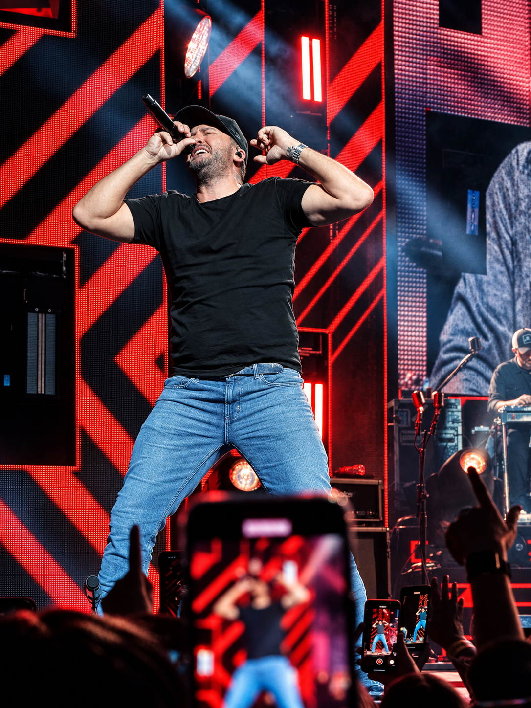 Male performer in black t-shirt and jeans energetically singing on a vibrant red and black concert stage.