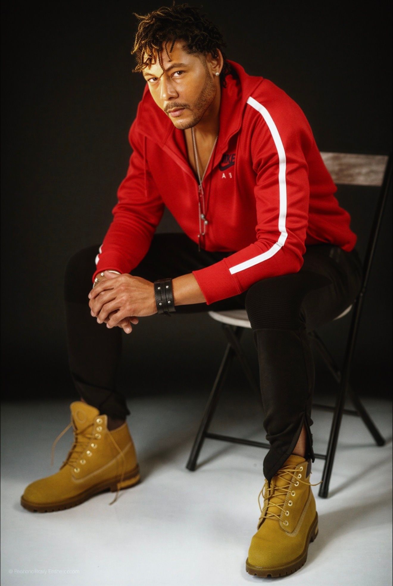 Man in red hoodie and tan Timberland boots sits confidently on a chair against a dark background.