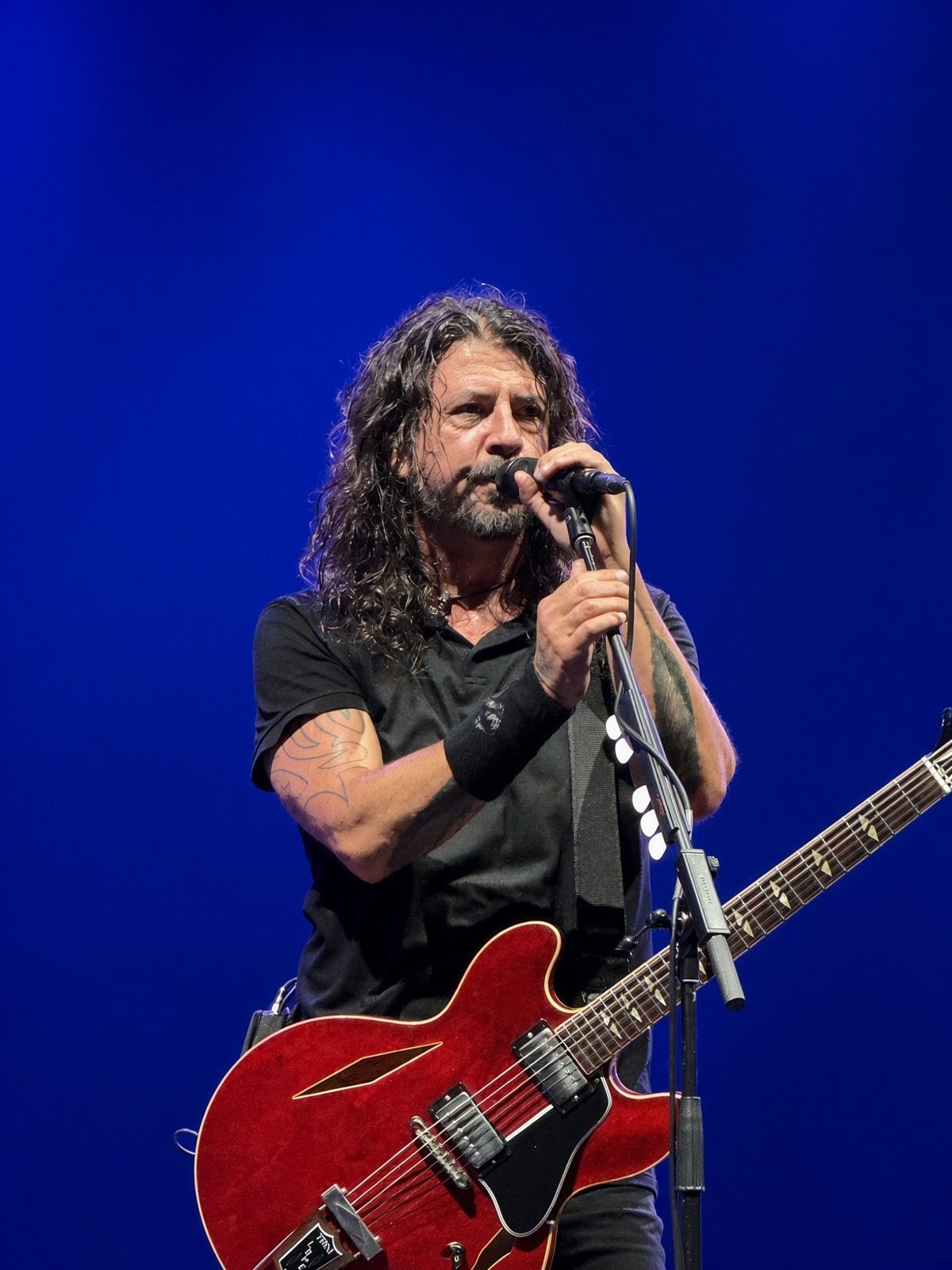 Male rock musician with curly hair singing into microphone while playing red semi-hollow electric guitar on blue-lit stage.