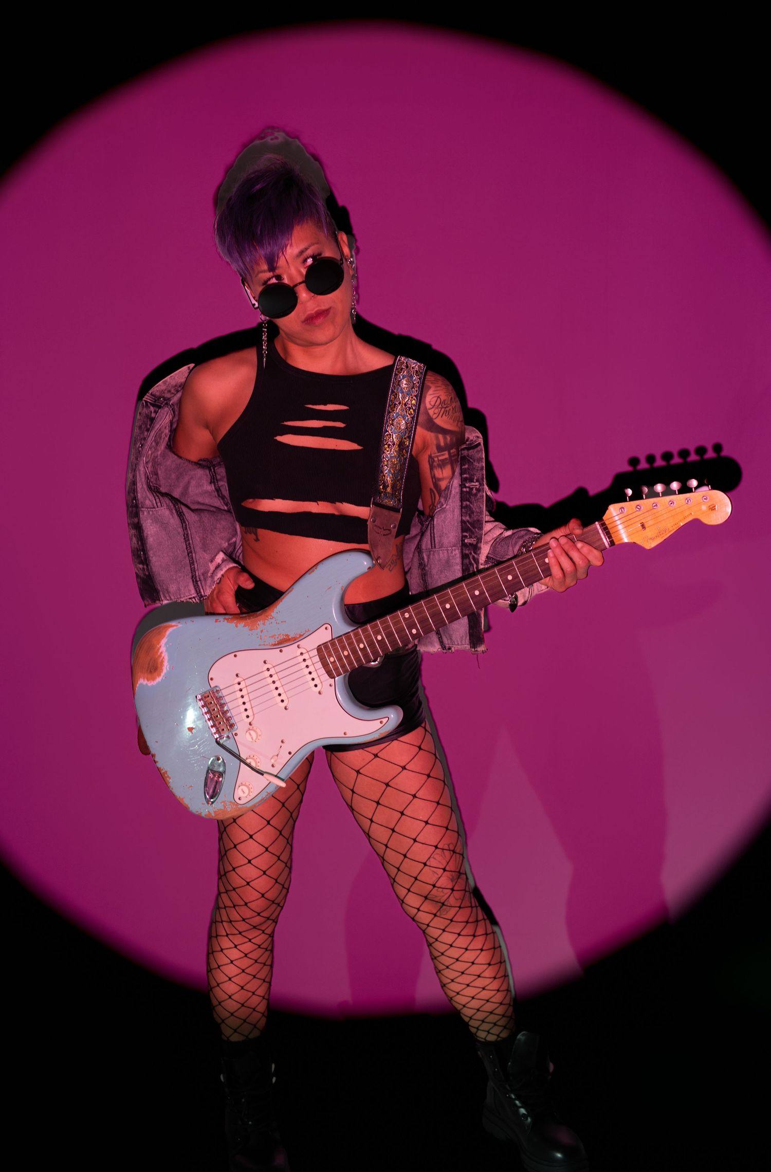 Female guitarist in fishnet tights and crop top plays blue Fender Stratocaster under pink spotlight.