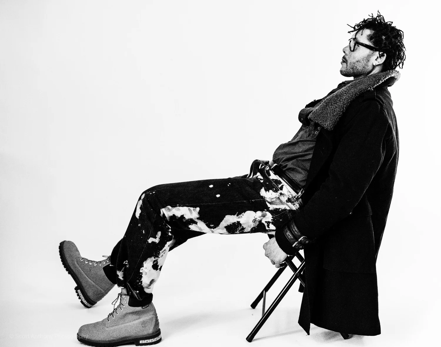 Black and white photo of a man in paint-splattered pants and boots, relaxing on a folding chair in a stylish pose.