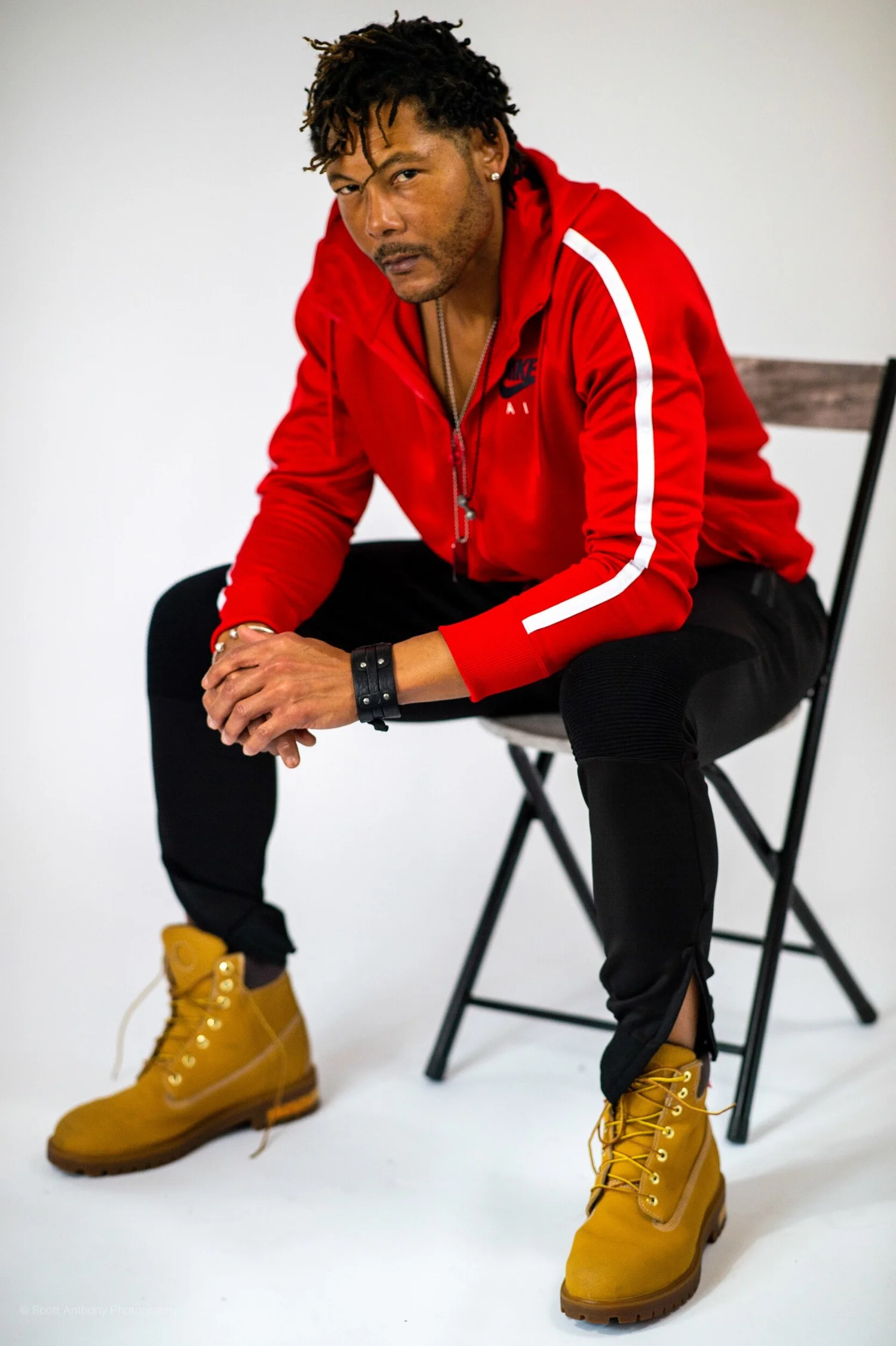 Man in red track jacket, black pants, and tan Timberland boots sits on a folding chair in a studio setting.